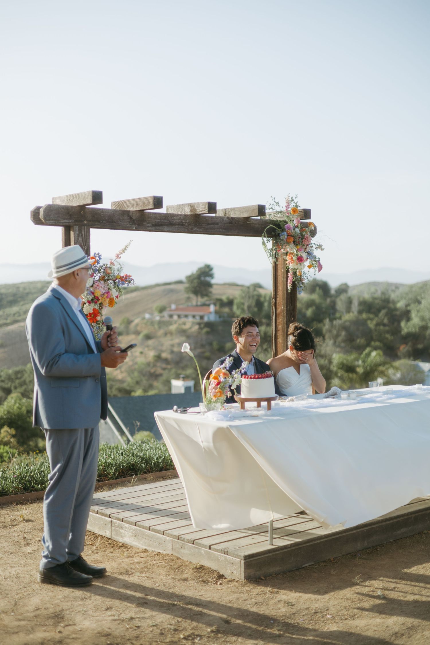 Outdoor wedding ceremony with couple at rustic wooden arch, floral arrangements, and rolling hills backdrop on a sunny day.