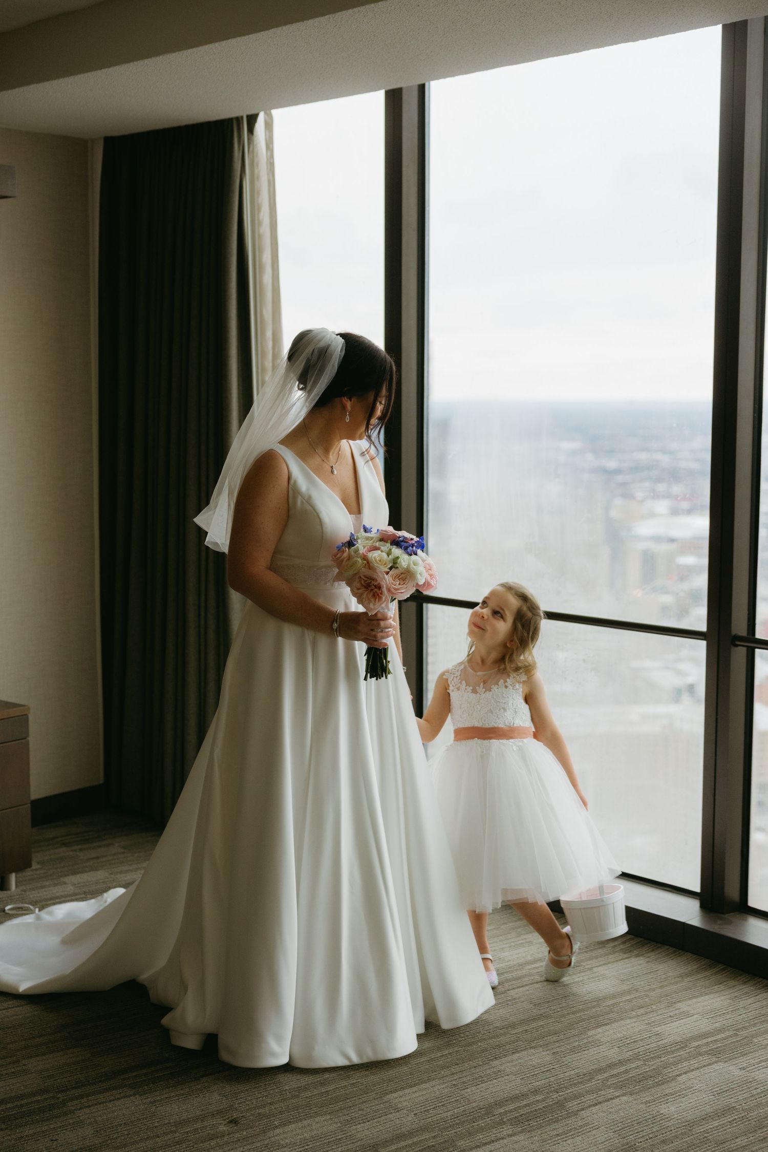 A tender moment between a bride in a white gown and a flower girl in a pink sash dress by large windows.