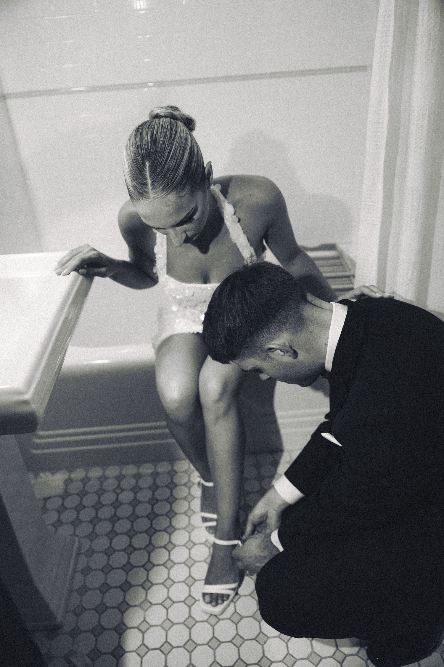 Black and white photo of a man in a suit fastening a woman's heels as she sits on a bathtub edge.