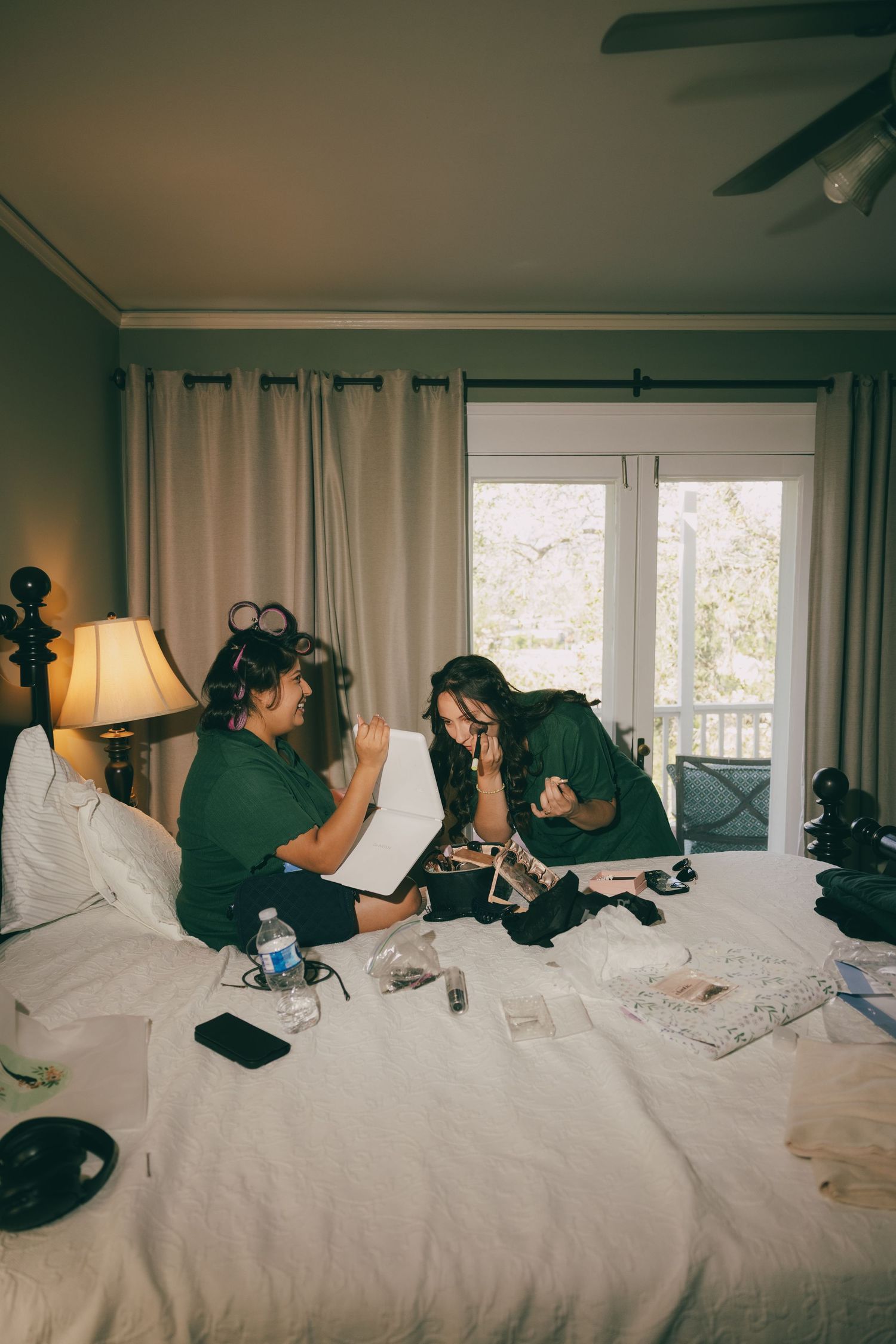 Two women in green shirts sit on a white bed reviewing photos in a warmly lit hotel room with curtains.