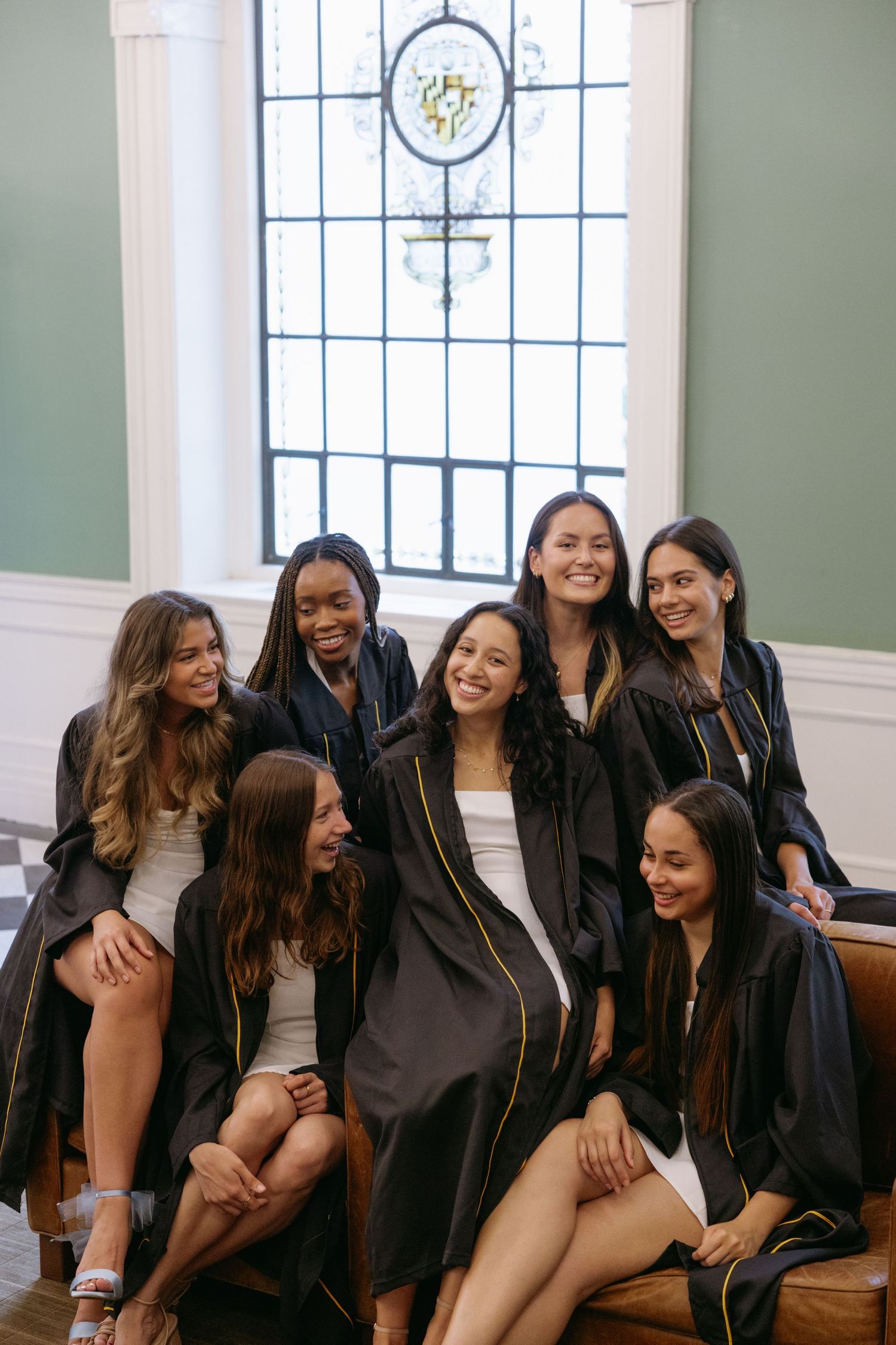 A group wearing matching black robes sit together on a wooden bench by a tall stained glass window.