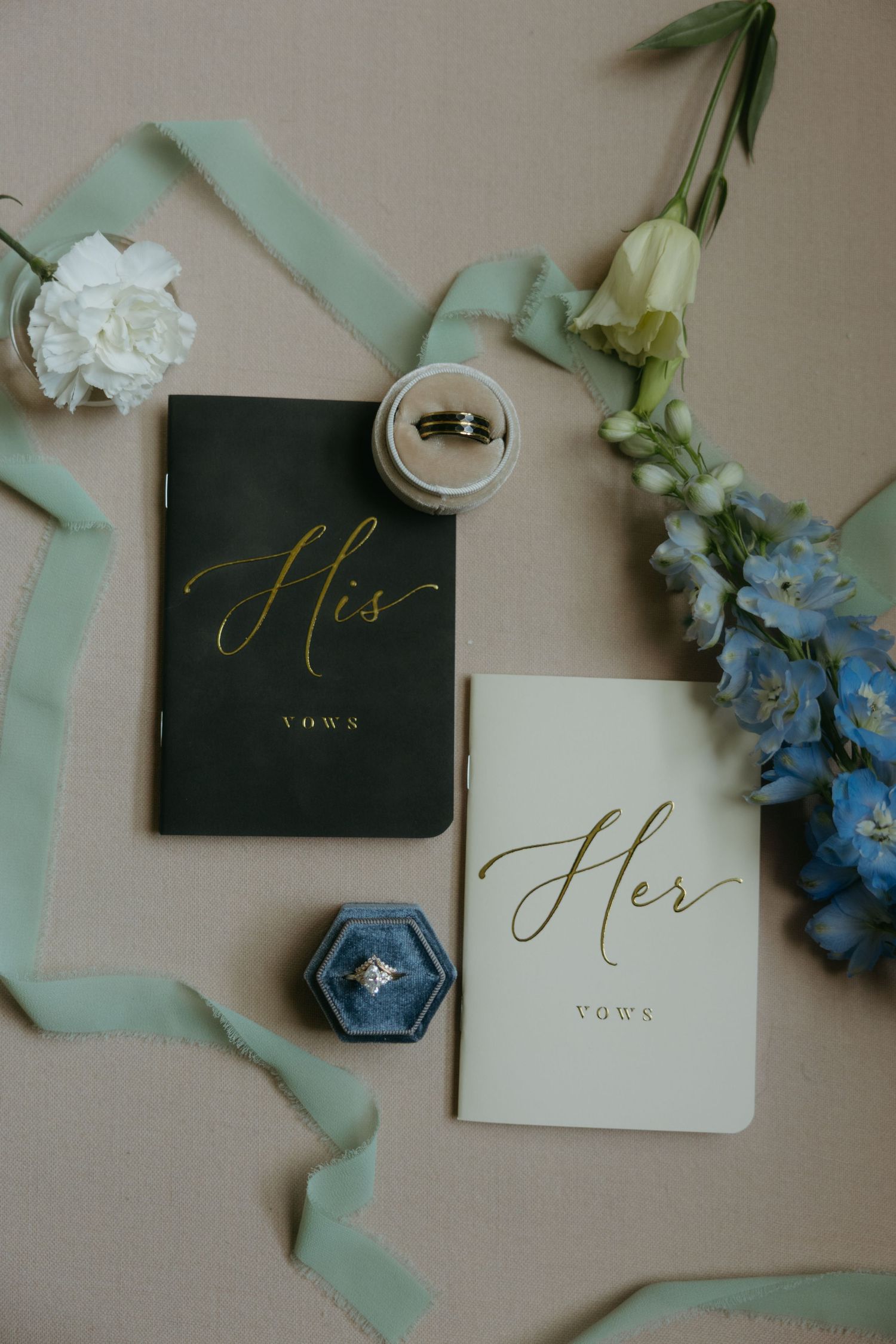 His and Her vow books with gold calligraphy, wedding rings, blue flowers, and mint ribbon on a neutral background.