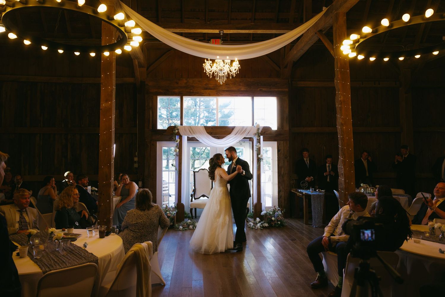 Couple shares first dance in rustic barn wedding venue with string lights, chandelier, and guests seated at decorated tables.