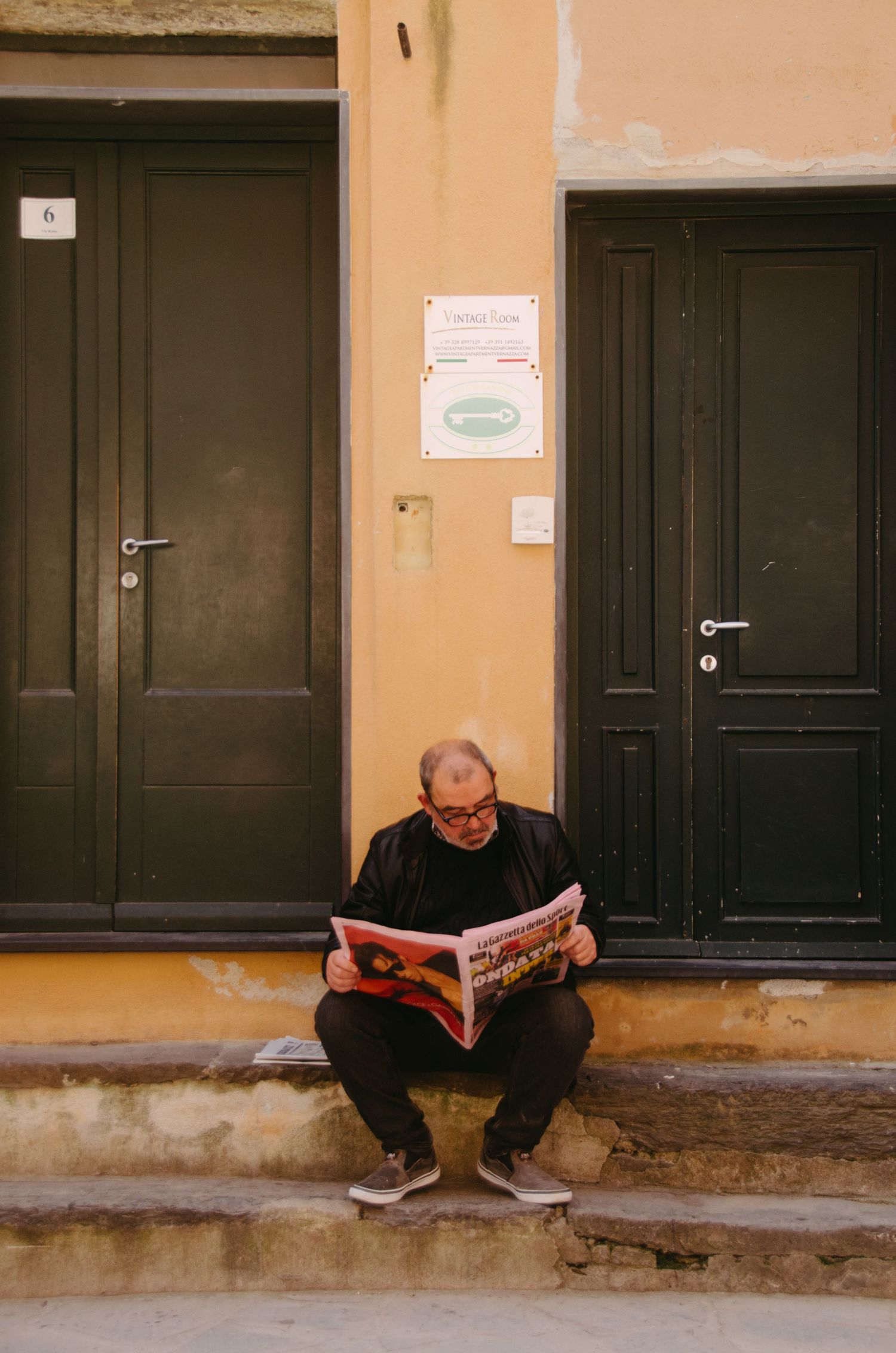Figure reading newspaper while sitting on steps between two dark doors in a yellow painted wall.