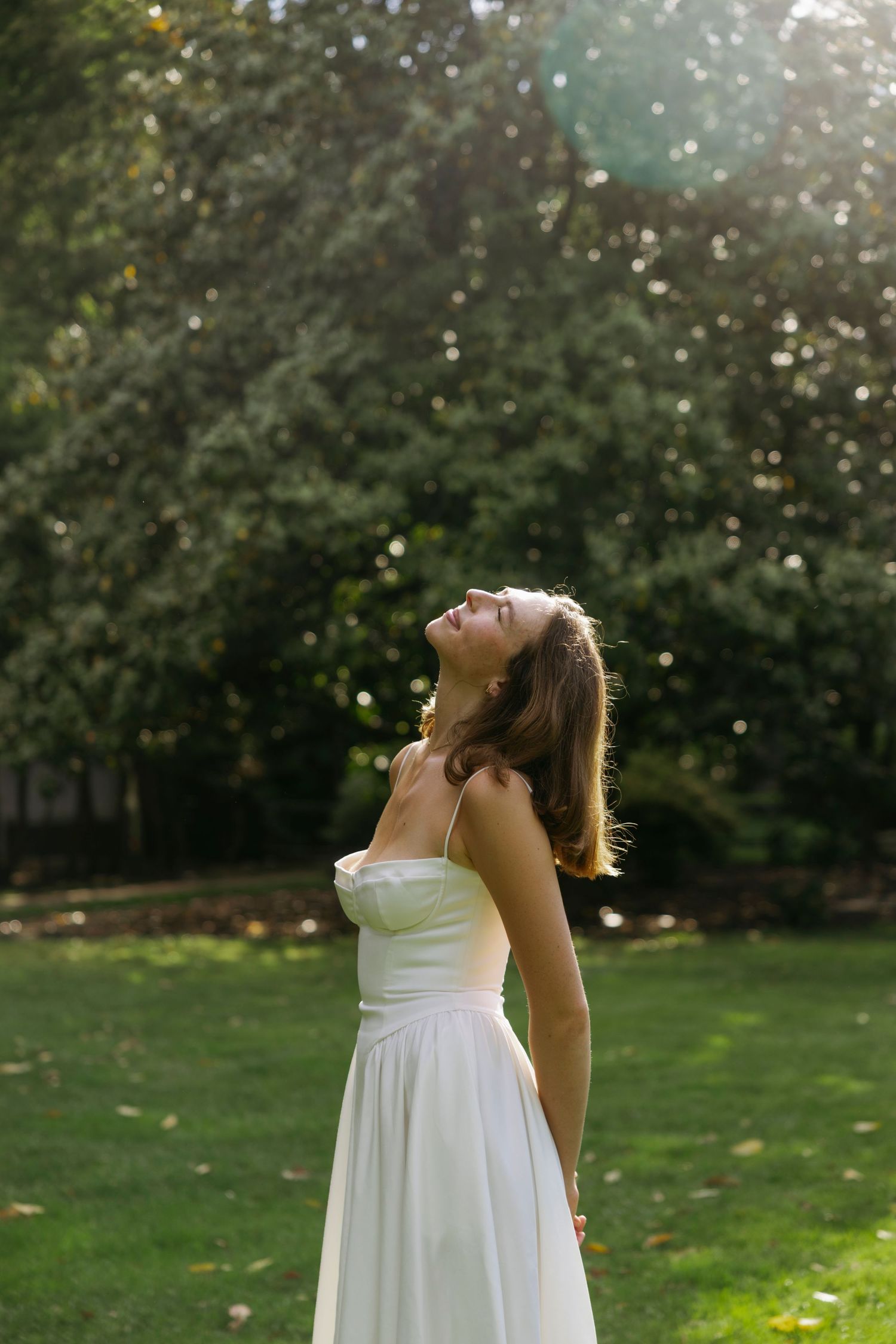 A person in a flowing white dress stands in a sunlit garden with their head tilted back amid green foliage.
