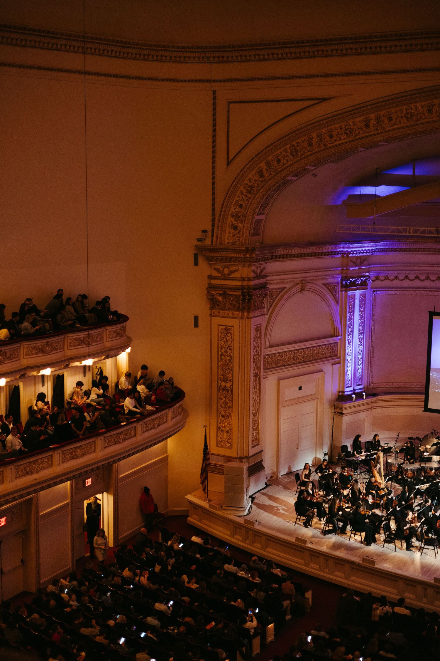 Orchestra performs on stage at Carnegie Hall while audience fills balcony seats in warm lighting with ornate architecture.