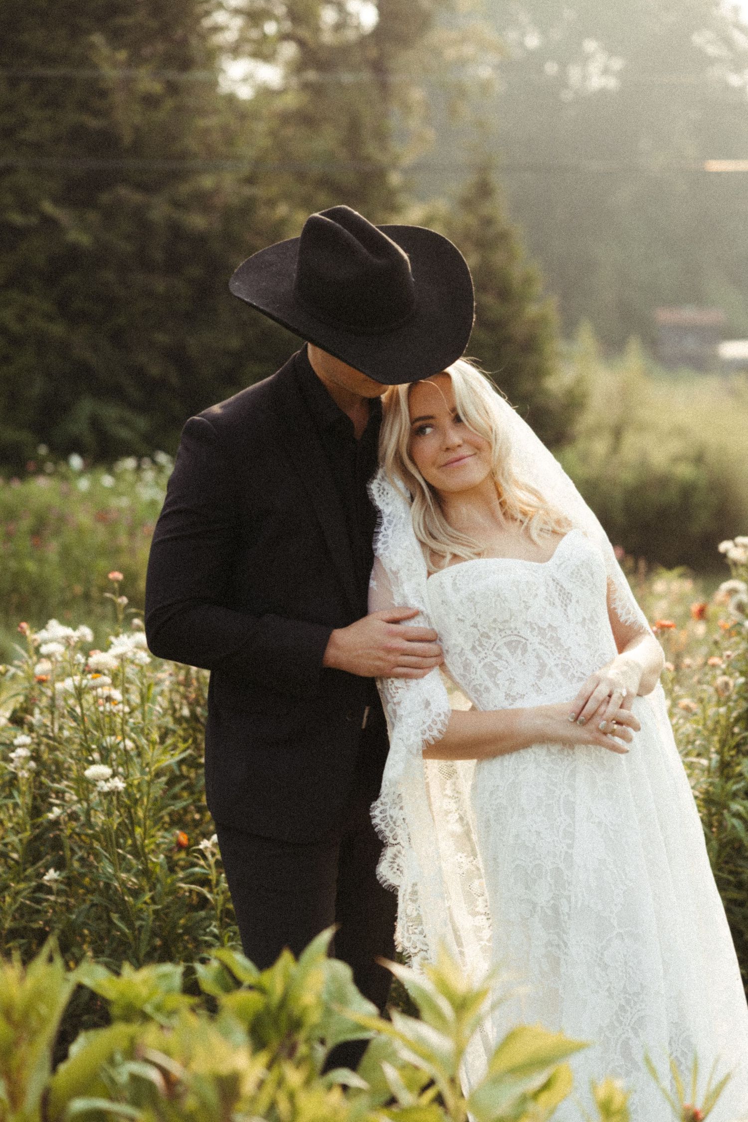 A couple in black and white wedding attire embrace in a sunlit garden during an intimate outdoor photo session.