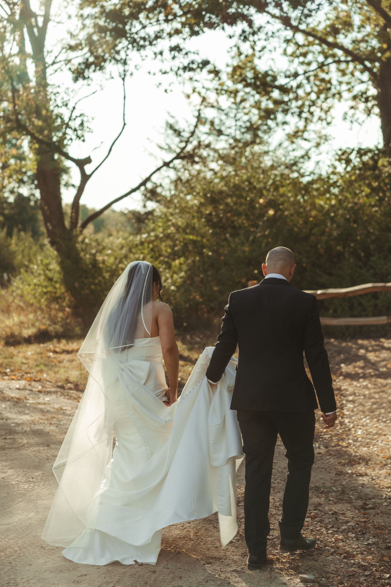 Newlywed couple walking together on a leaf-covered path through autumn trees with bride's veil flowing in the breeze.