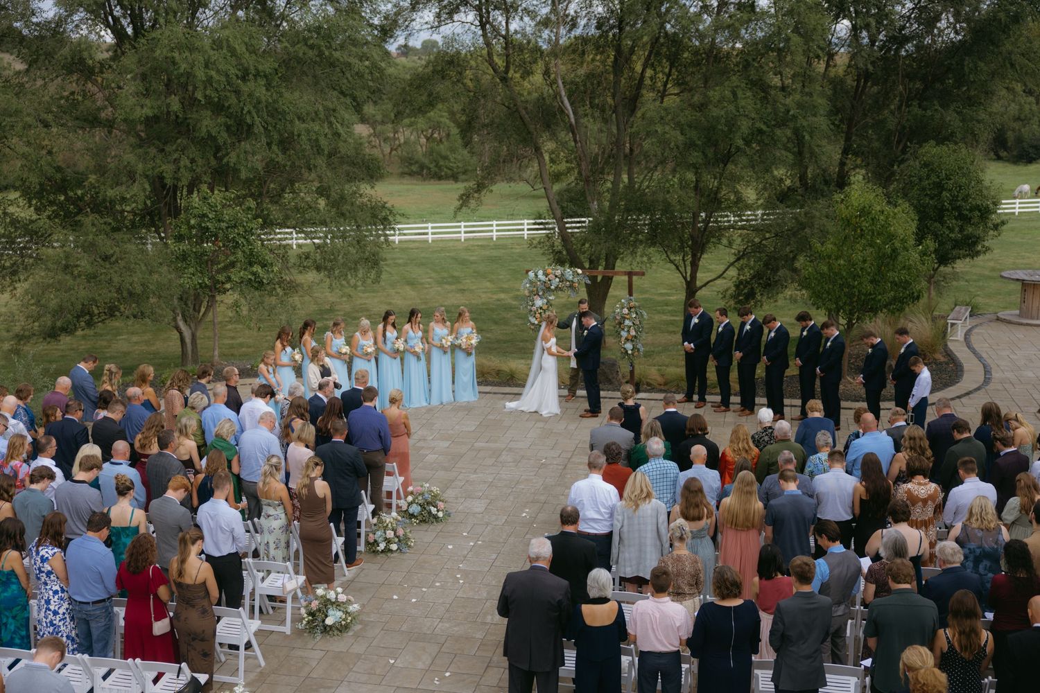 Outdoor wedding ceremony gathering with guests standing in rows on grassy lawn surrounded by trees and white fencing.