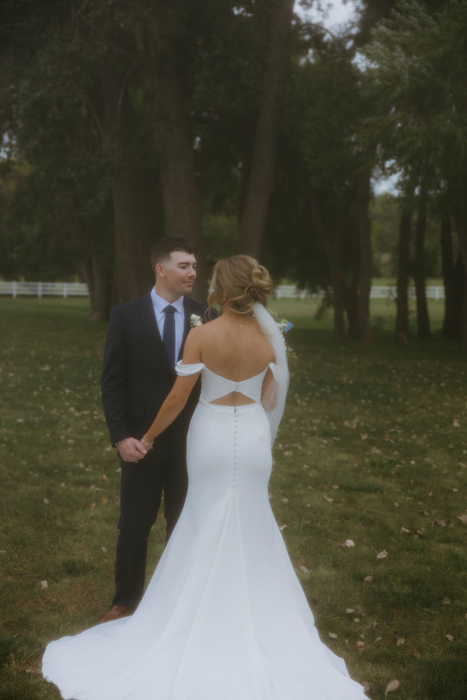 A bride in a flowing white ballgown dress stands in a wooded outdoor setting during a wedding ceremony.