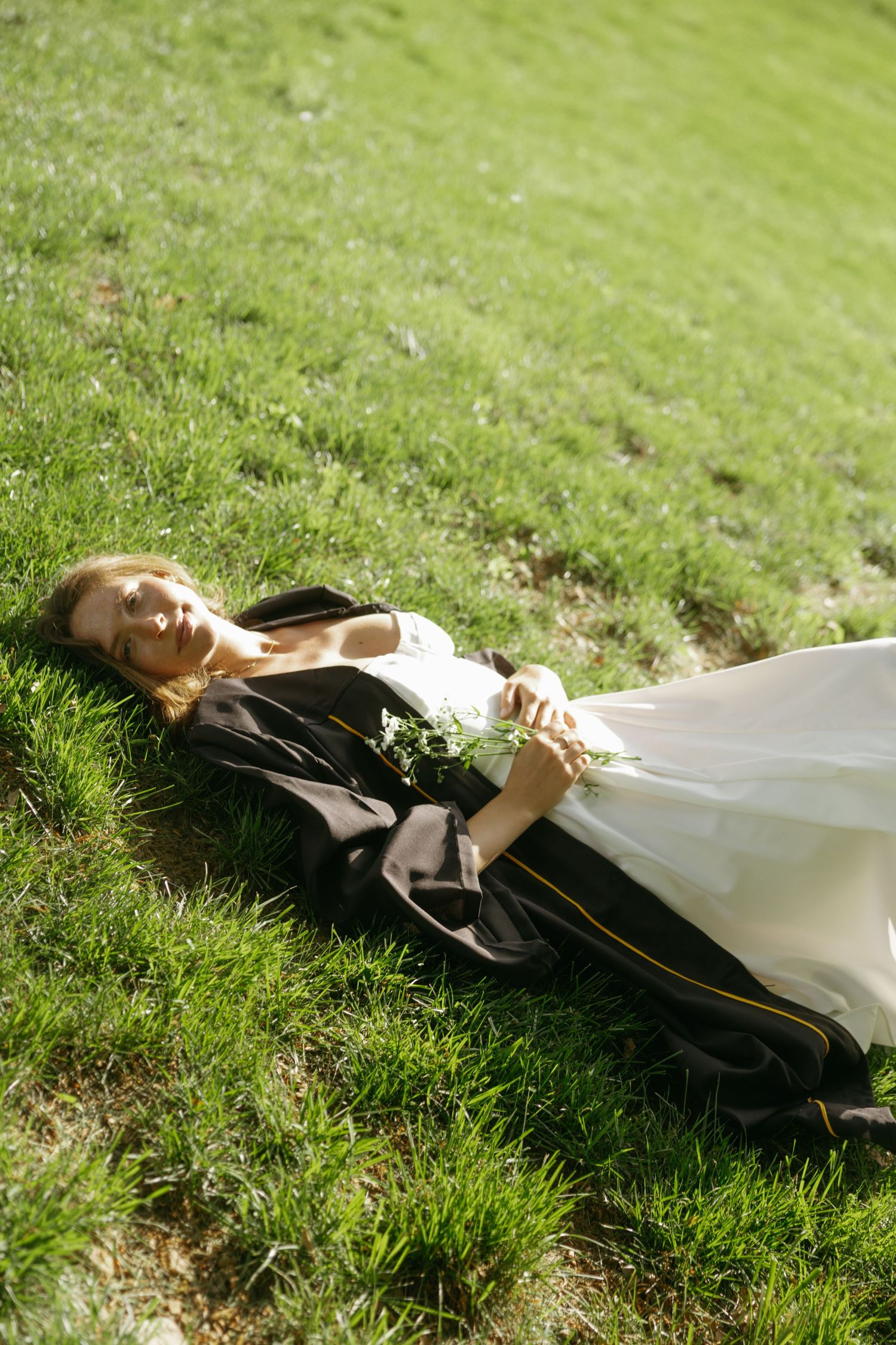 A figure wearing a dark jacket and white dress reclines peacefully on lush green grass in sunlight.