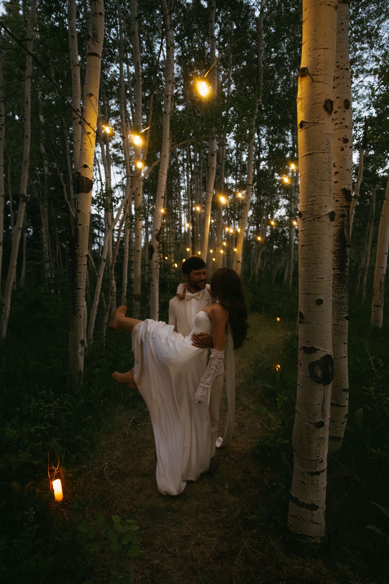 Couple in white attire dancing among lit birch trees at dusk with string lights glowing in an enchanted forest.