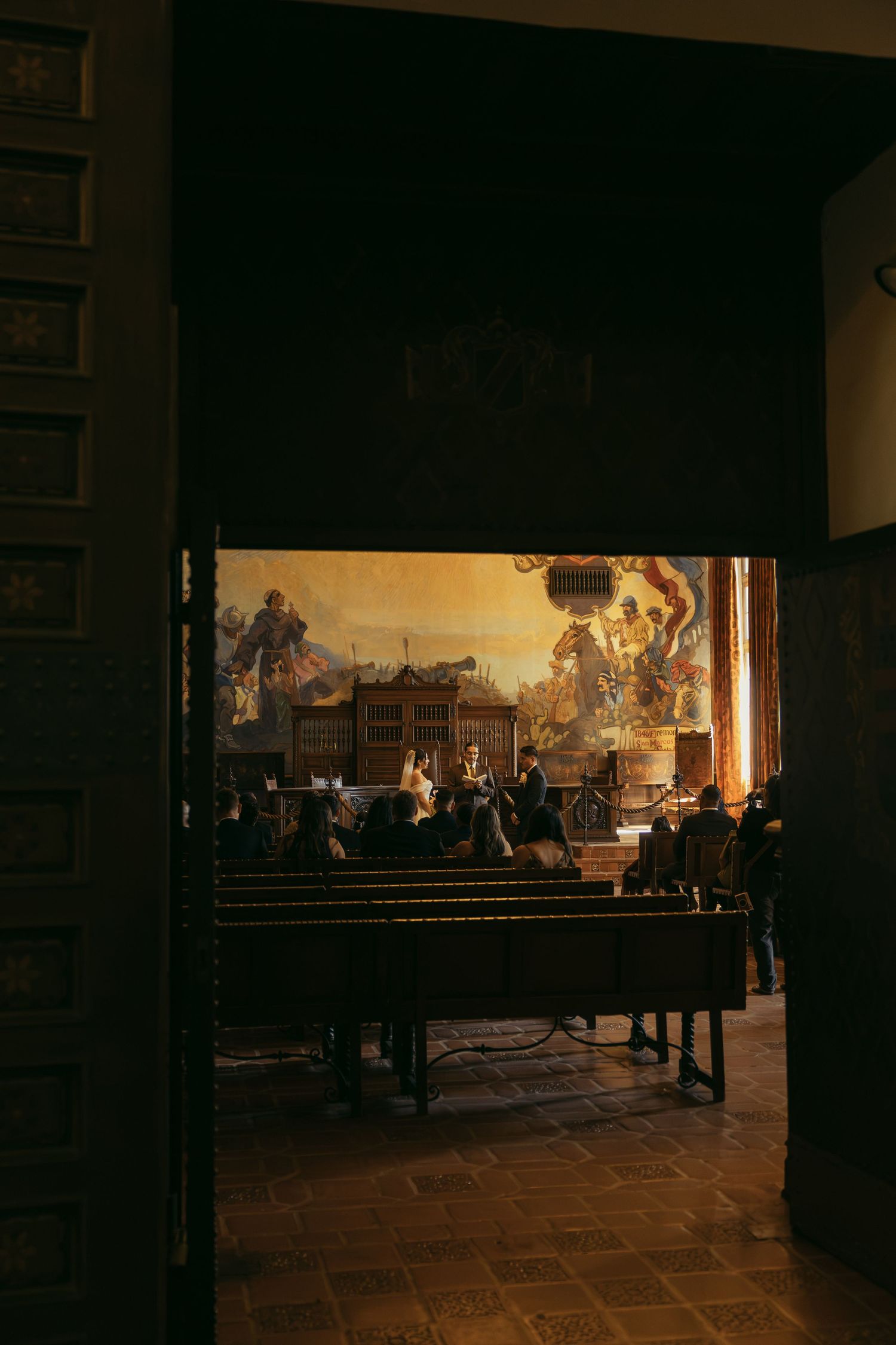 Dark doorway reveals musicians performing before a grand Renaissance mural painting in an ornate historic hall.