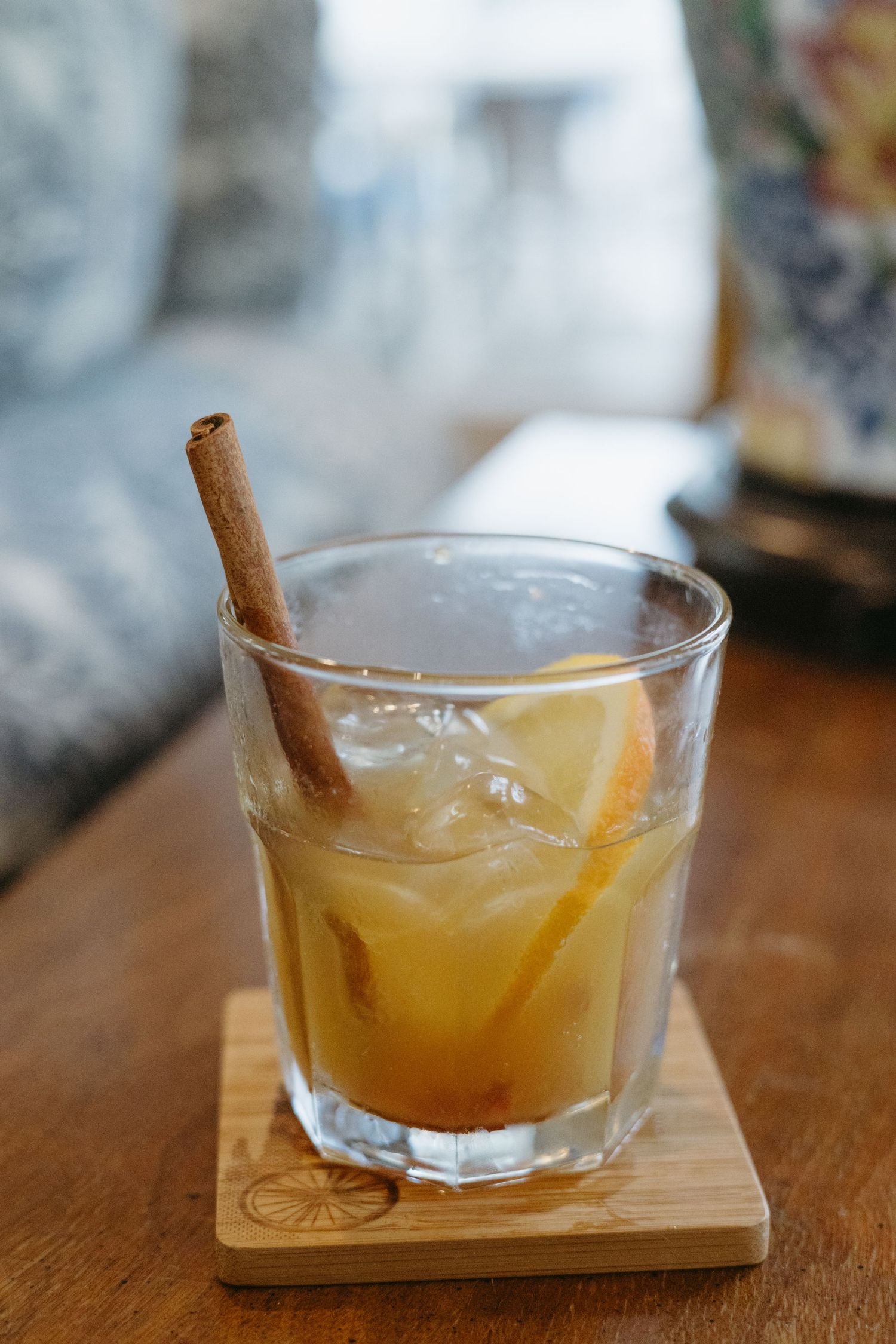 A cocktail glass filled with amber liquid garnished with a cinnamon stick and citrus slice sits on a wooden coaster.