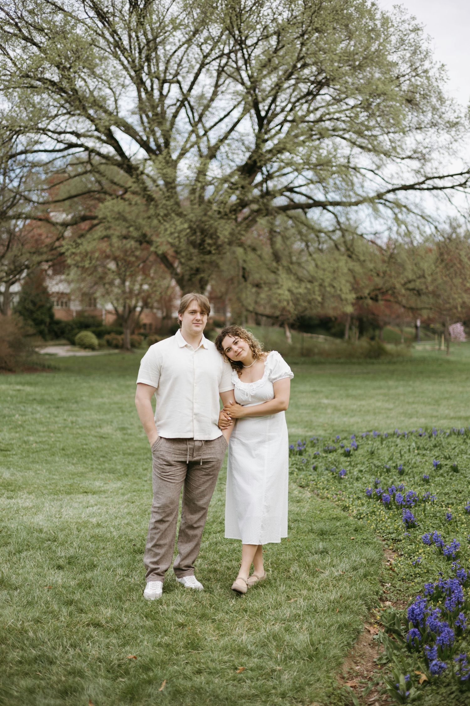 A couple stands together on a green lawn with blooming purple flowers and a large tree in the background.