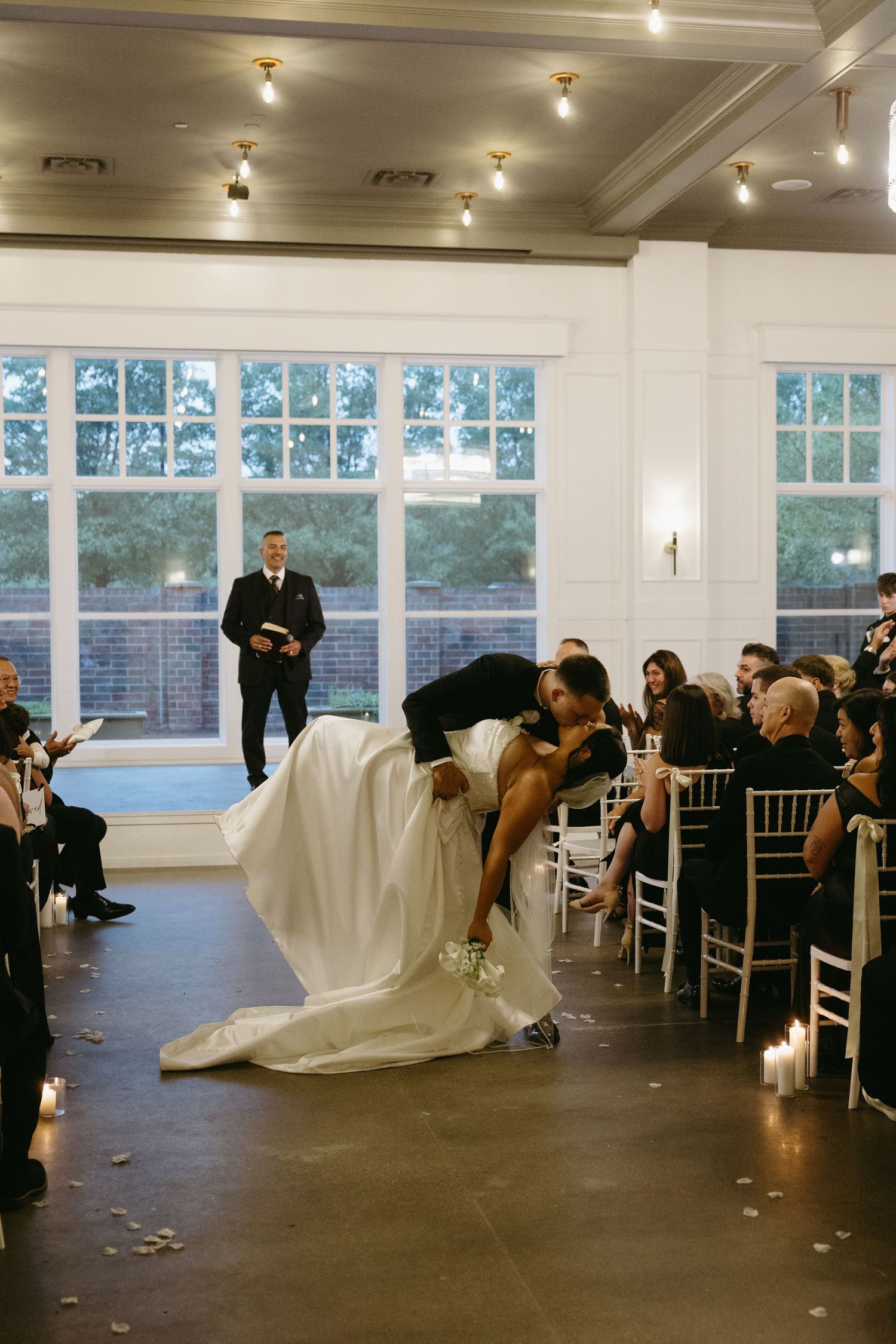 Bride and groom share a romantic dip kiss during their indoor wedding ceremony while guests watch from white chairs.