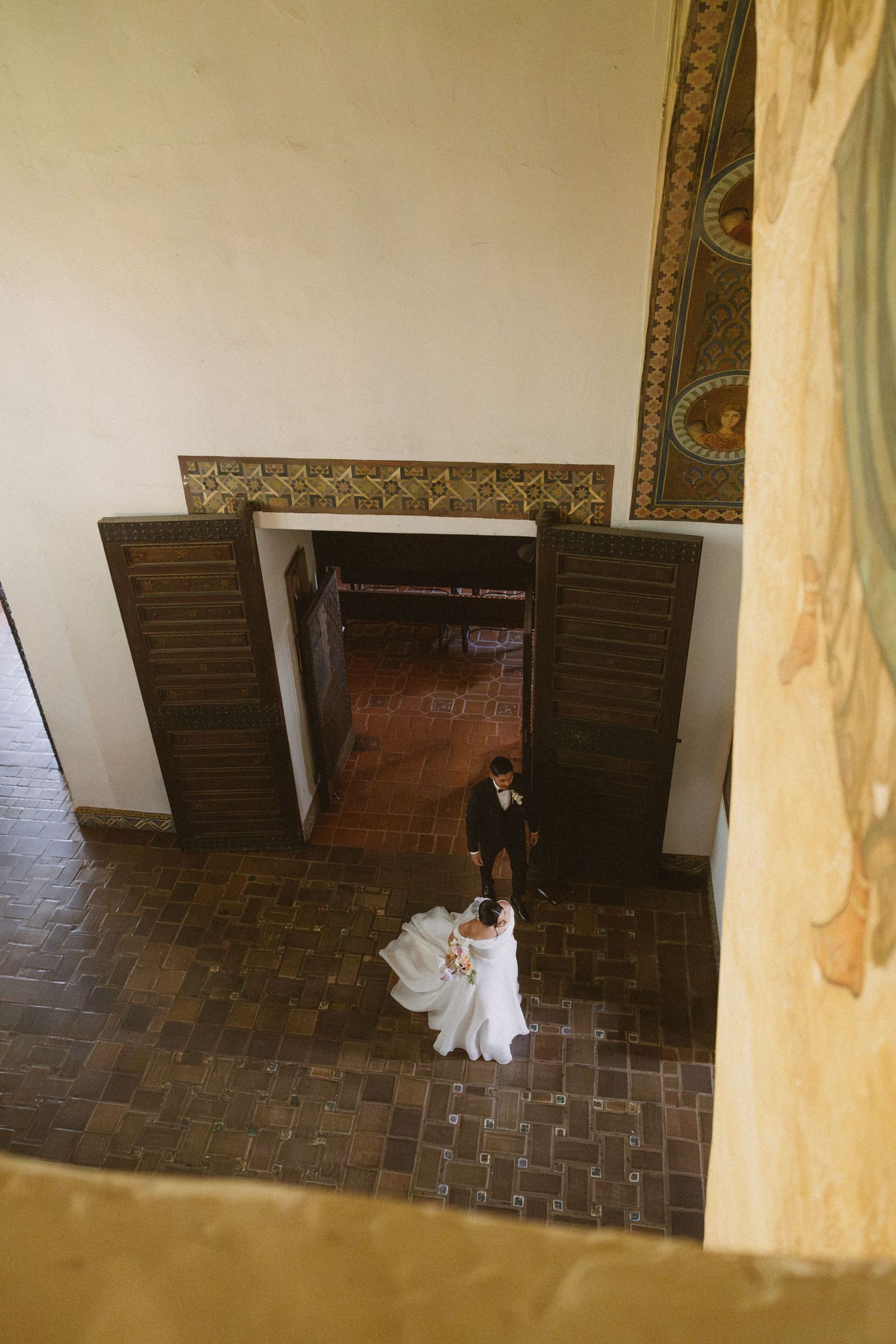 Aerial view of bride and groom entering ornate venue through wooden doors with decorative tile floors.