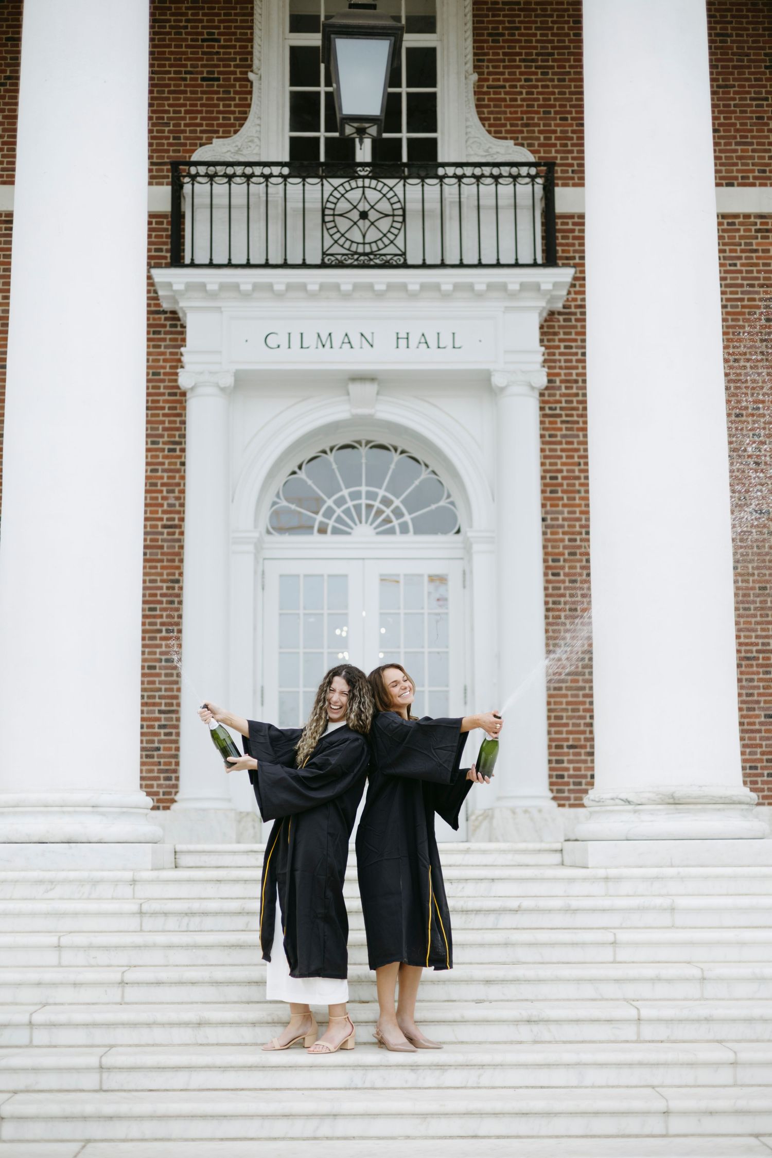Two graduates in black caps and gowns celebrate on white steps in front of historic Gilman Hall's grand entrance.