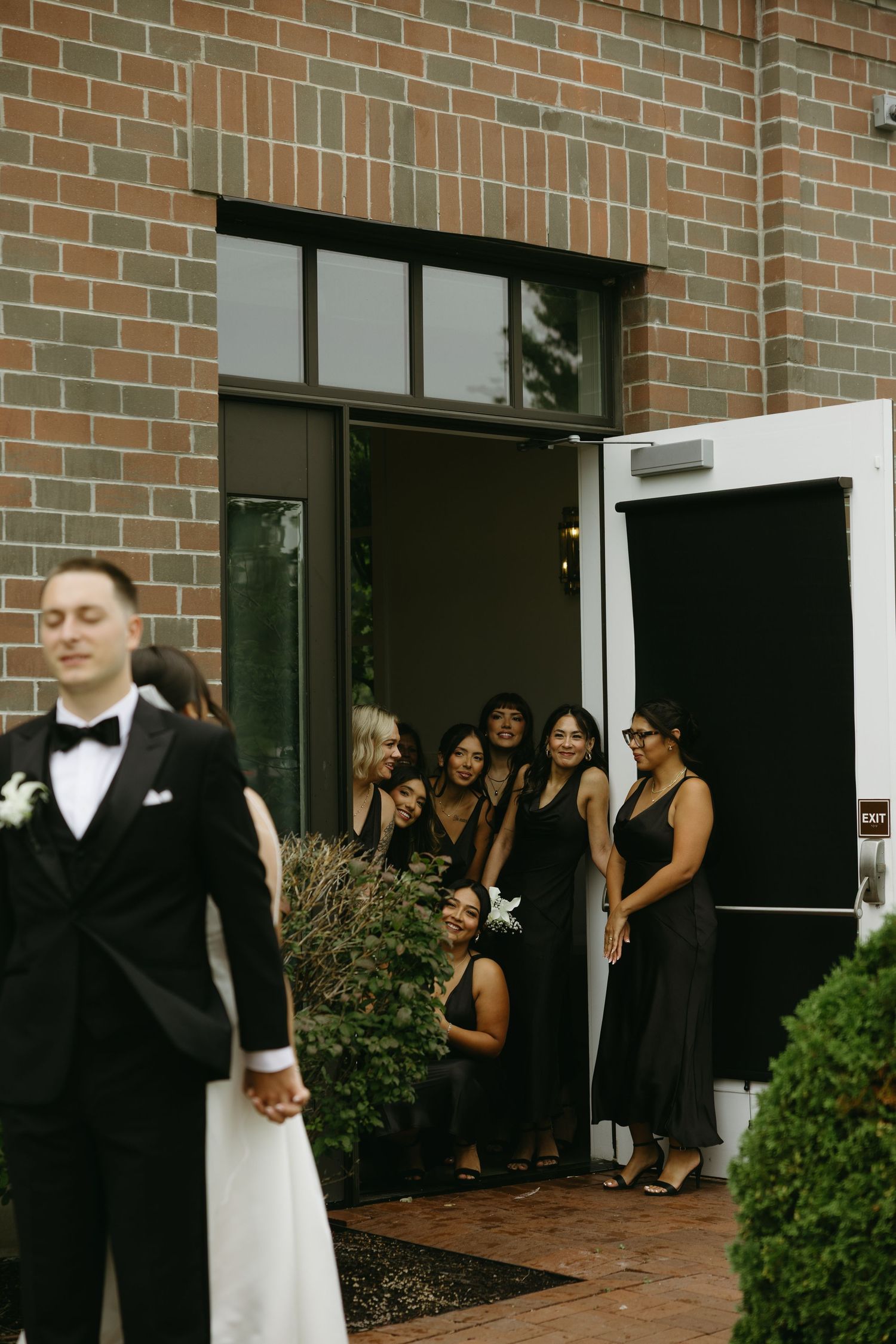 Bridesmaids in black dresses peek from doorway as groom in tuxedo stands outside brick building.