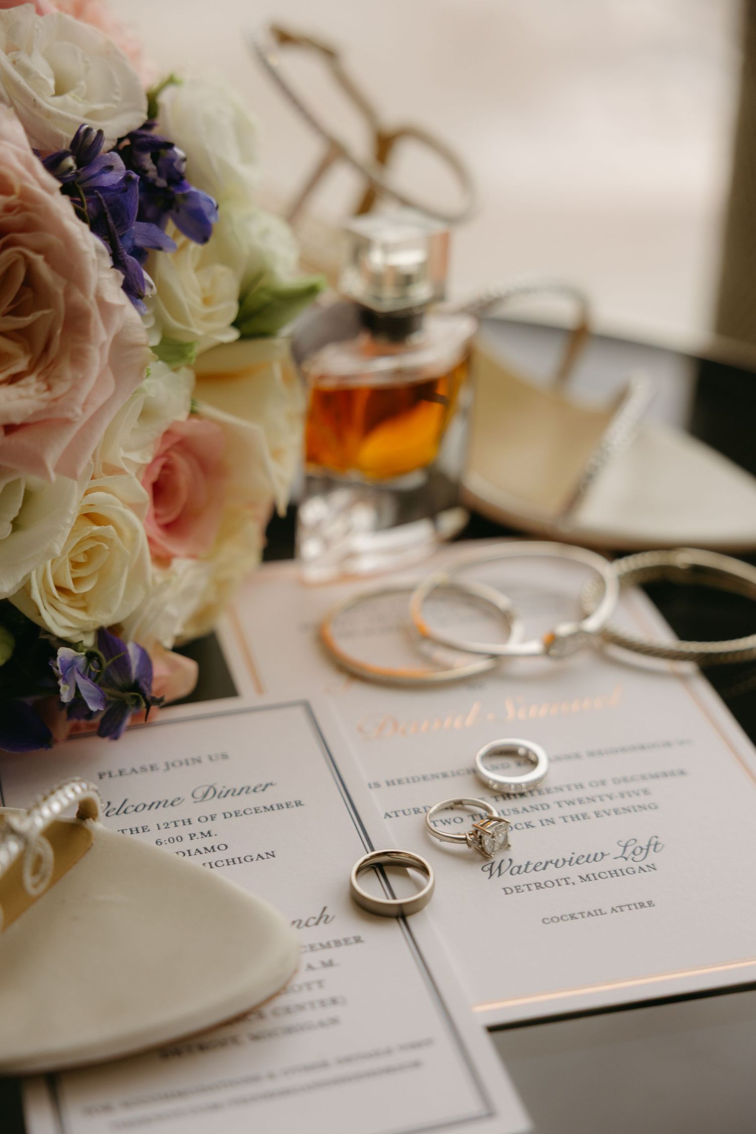 Wedding rings rest elegantly on white invitation cards with delicate pink and purple floral arrangements nearby.
