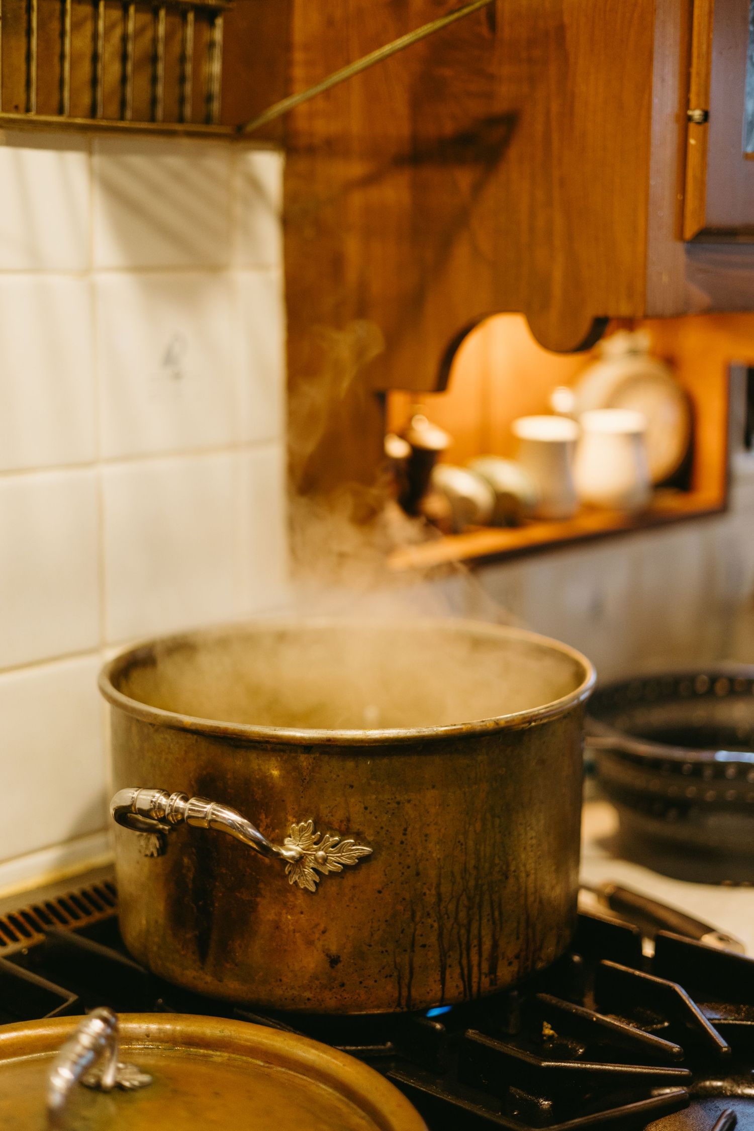 Steam rises from a tarnished copper pot with butterfly handles simmering on a stovetop against a wooden kitchen interior.