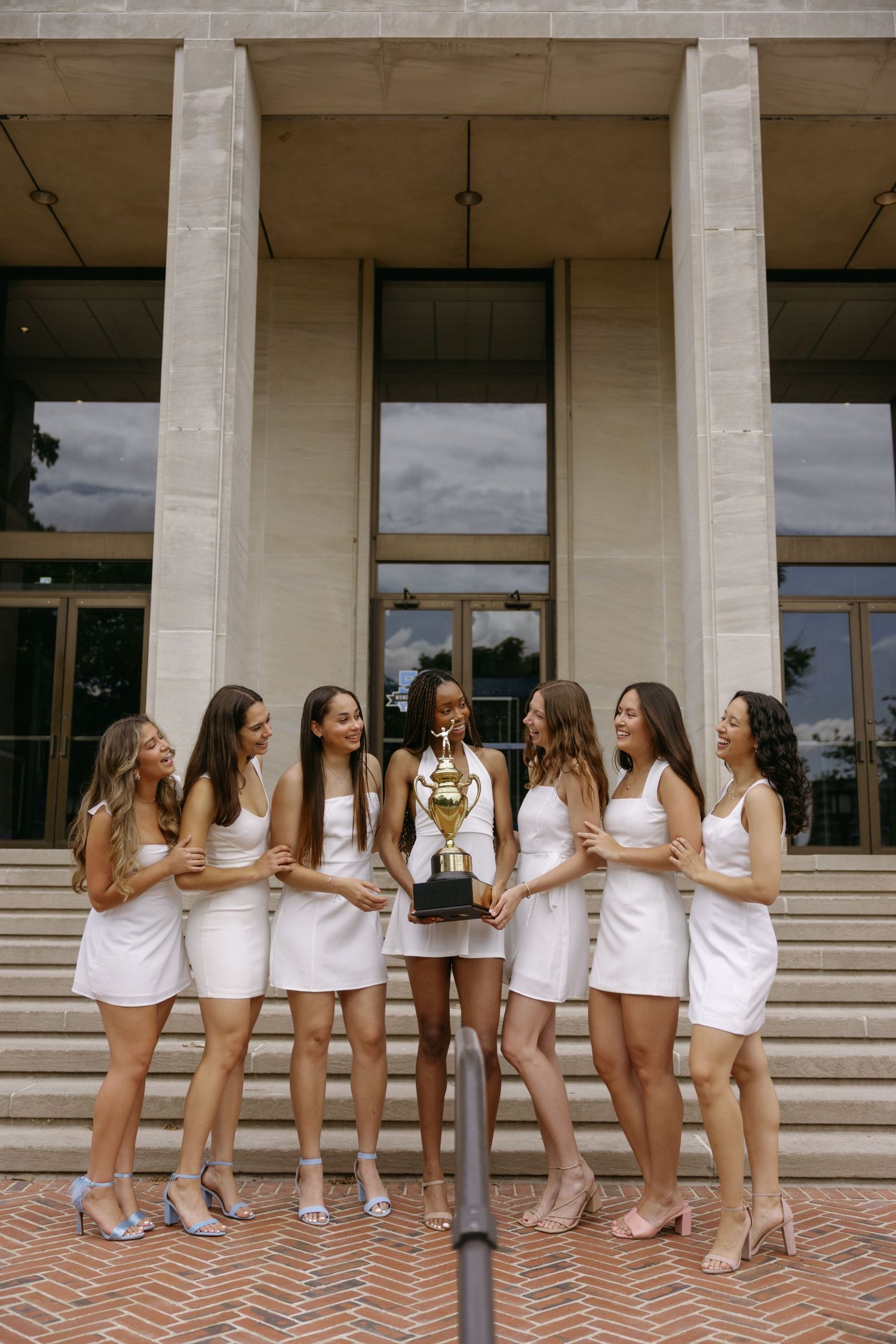 A group in matching white outfits stands in a celebratory pose on brick steps of a building with columns.