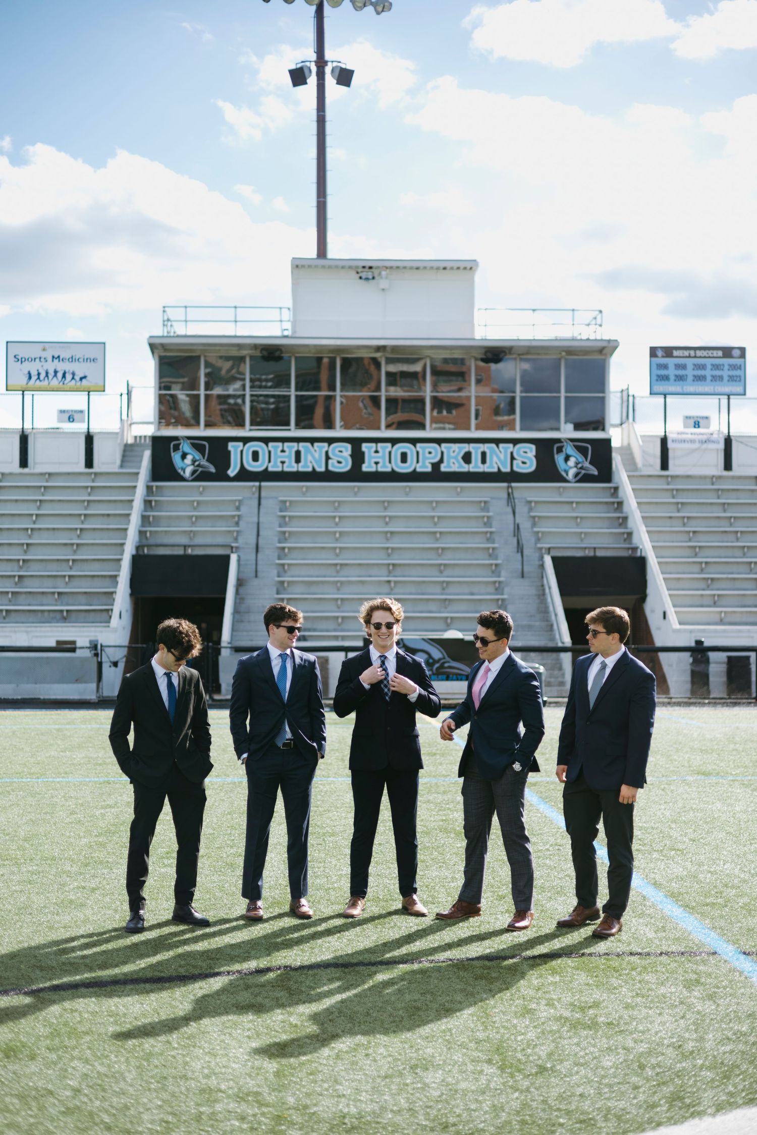 A group of well-dressed men in suits stand on a football field in front of stadium bleachers and a press box.