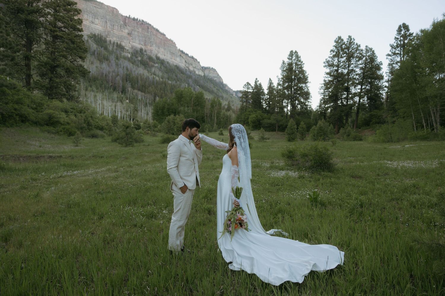 A bride and groom share a romantic moment in a lush green mountain meadow, surrounded by tall pines and rocky cliffs.