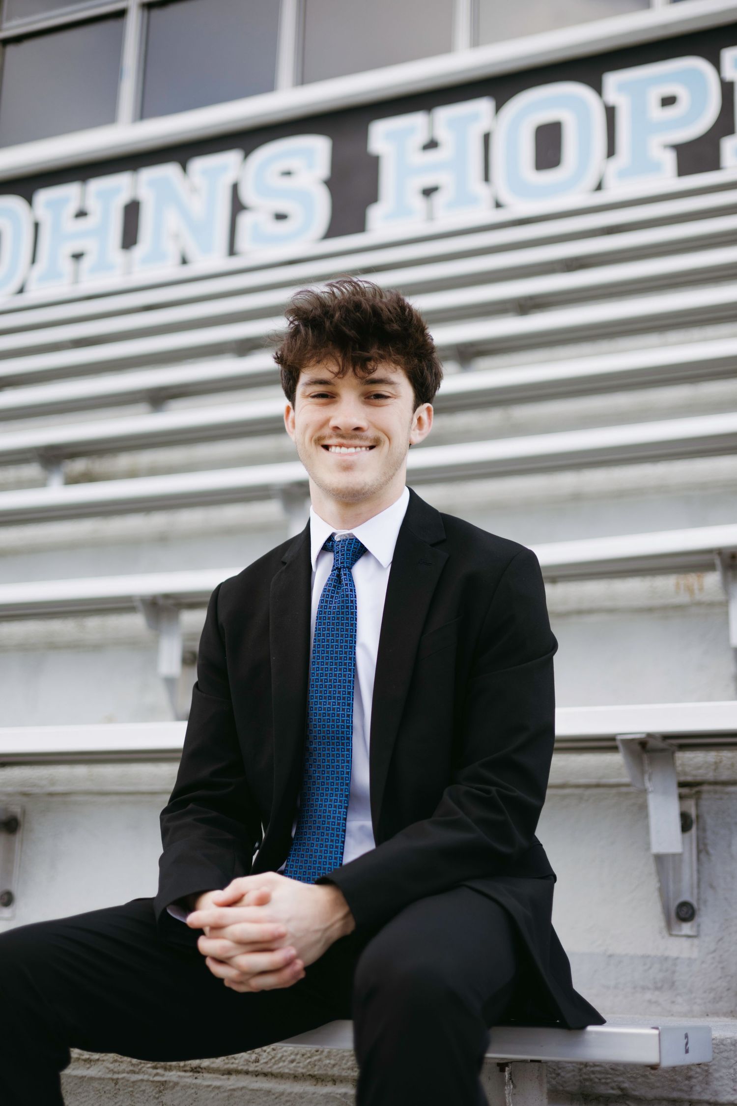Young professional in dark suit and blue tie sits smiling on stairs in front of Johns Hopkins building sign.