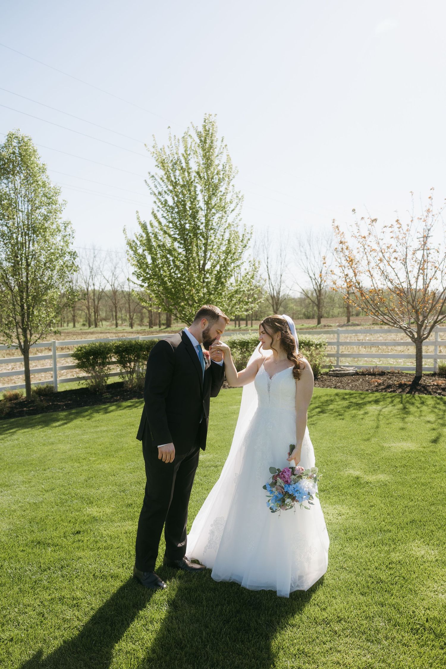 Groom kissing bride's hand on lush green lawn with white fence and spring trees in background on sunny wedding day.