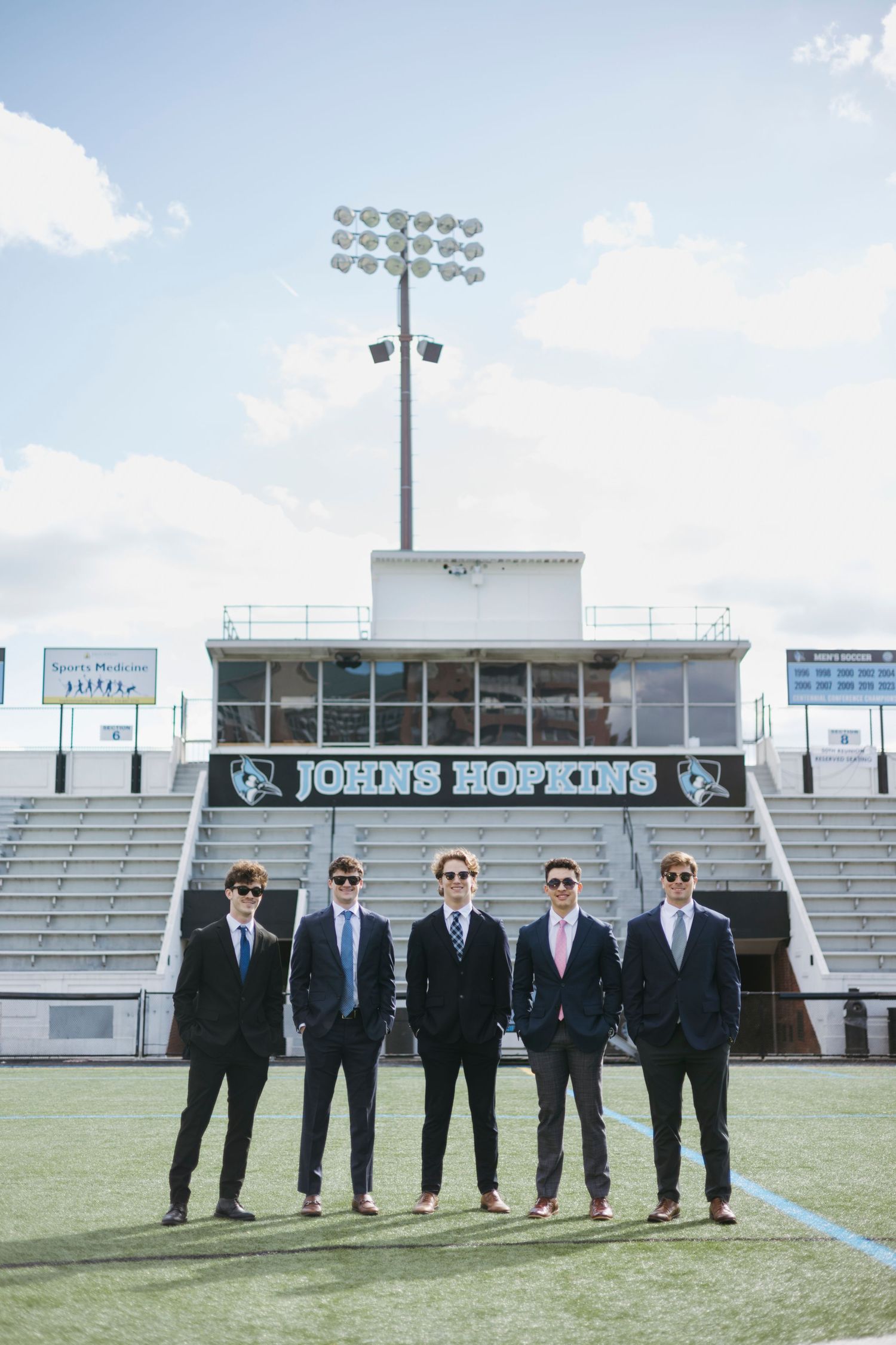 Five people in dark suits stand in formation on a football field in front of stadium bleachers with 'JOHNS HOPKINS' signage.
