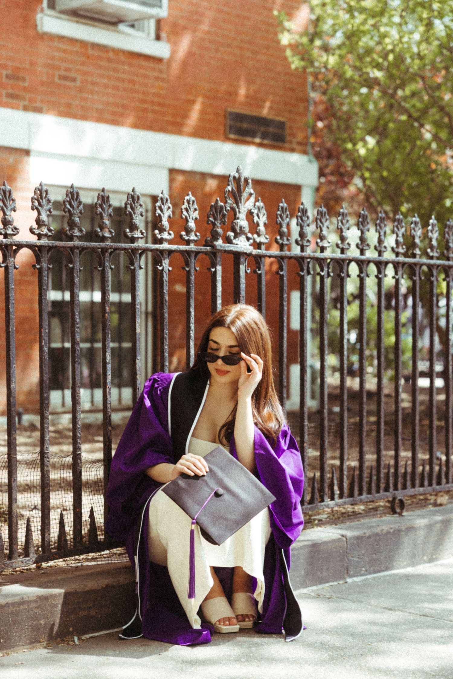 Fashionable woman in purple coat sits on steps near ornate iron fence while reading on sunny day.