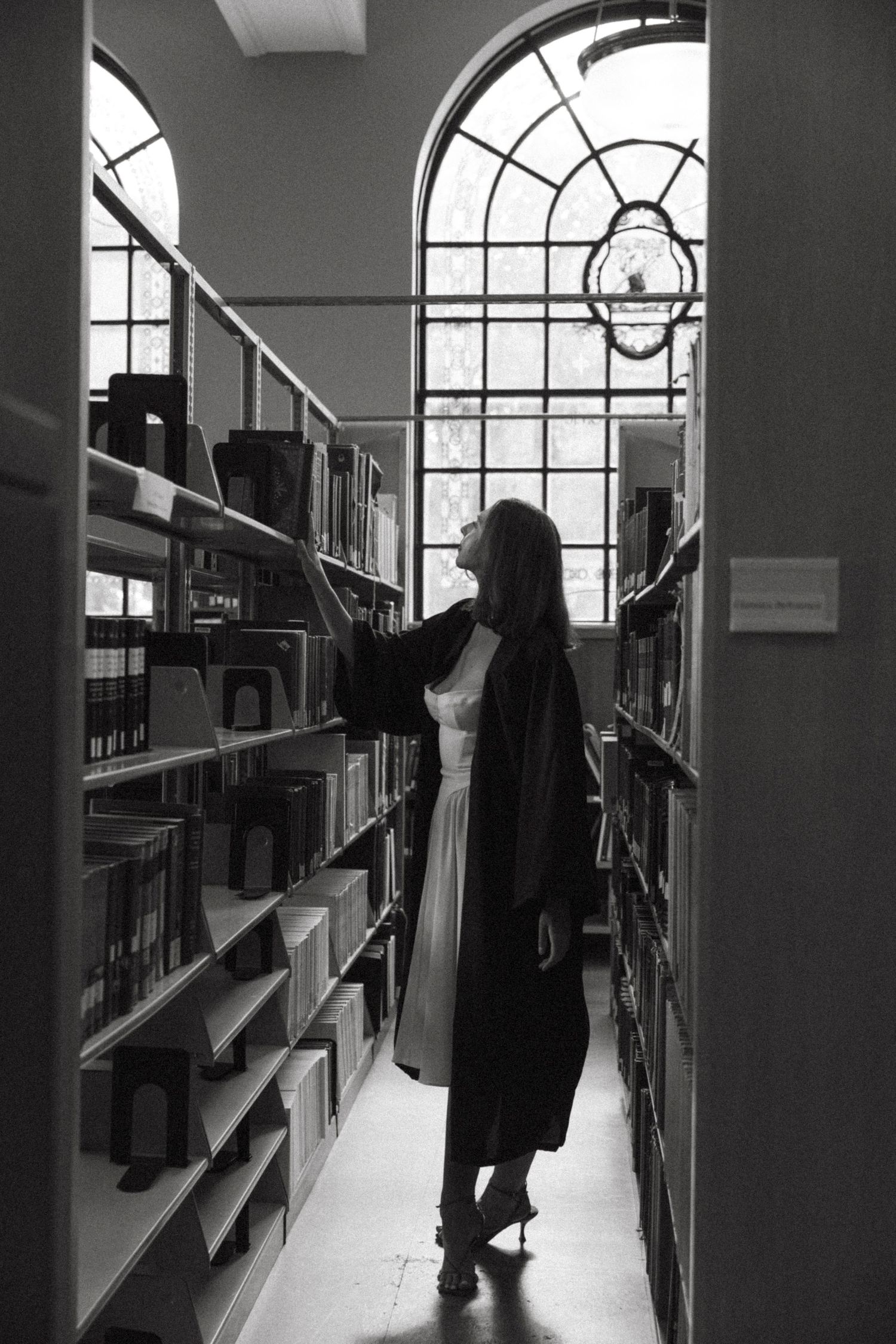 A silhouetted figure browses bookshelves in a library with large arched windows and natural light streaming in.