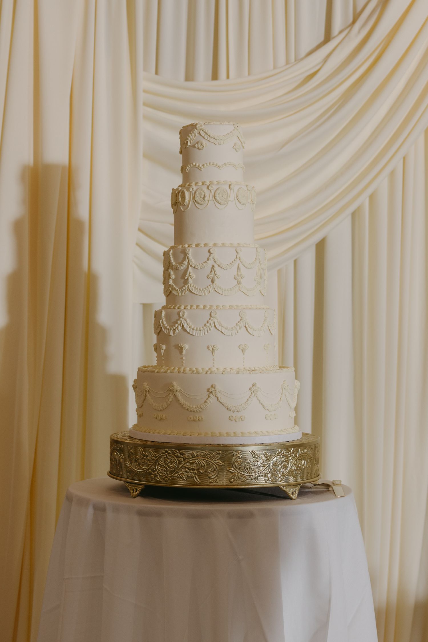 Elegant five-tier white wedding cake with gold detailing on ornate stand against draped cream backdrop.