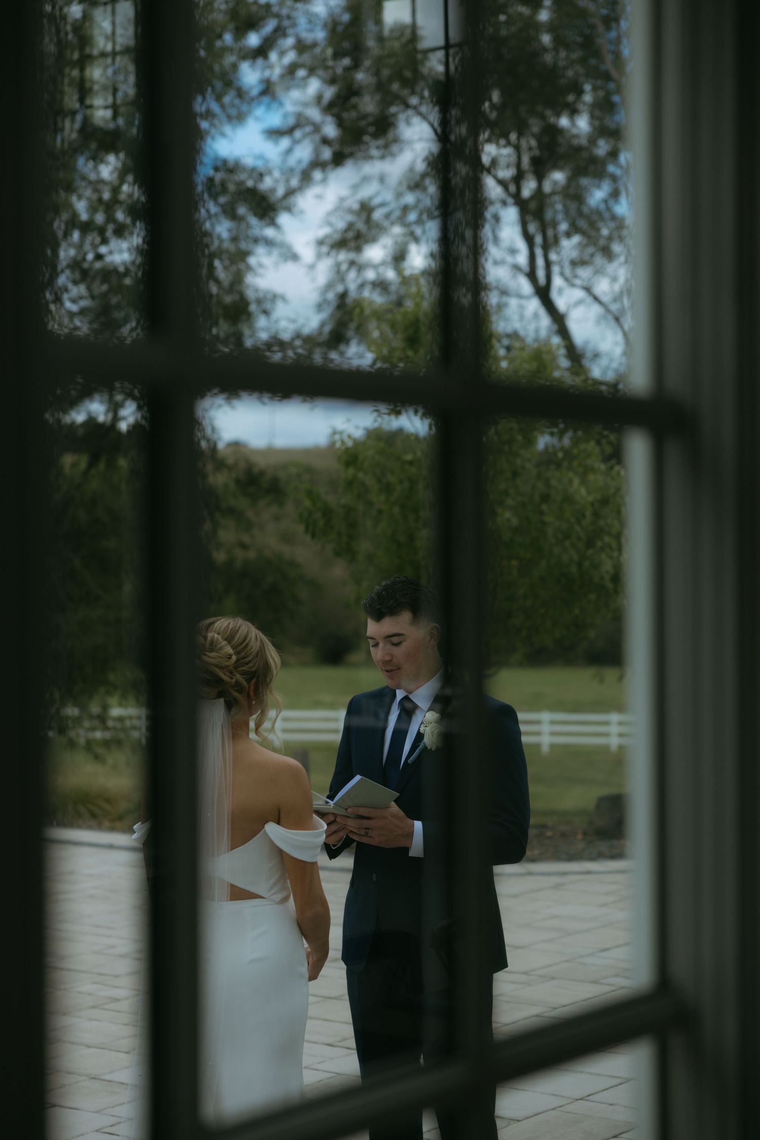 A couple stands on an outdoor patio surrounded by glass windows and white fencing in a scenic countryside setting.