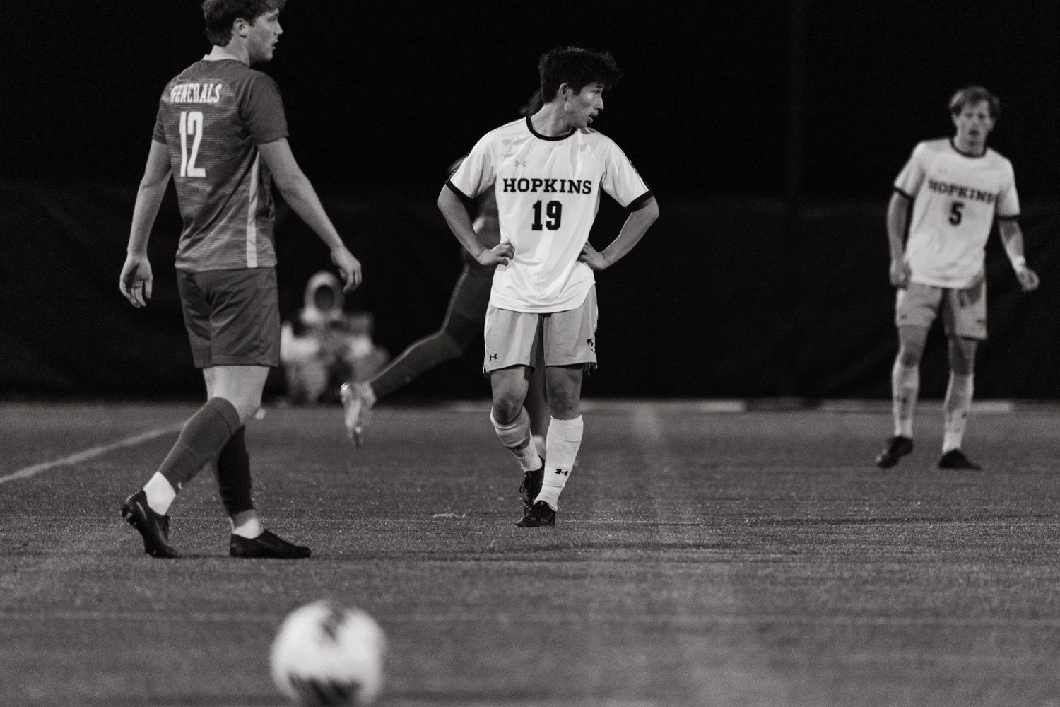 Soccer players on a field at night with a ball in the foreground during a competitive match.