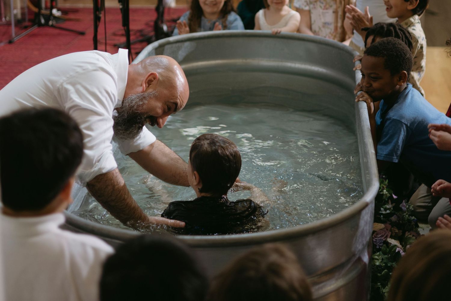 A religious baptism ceremony takes place as a person is lowered into a large metal tub of water surrounded by observers.