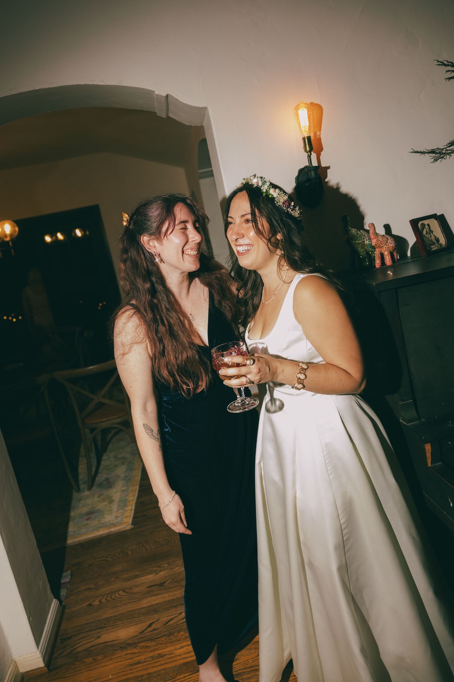 Two women smiling and laughing together at a wedding celebration, one wearing a white bridal gown with floral crown.