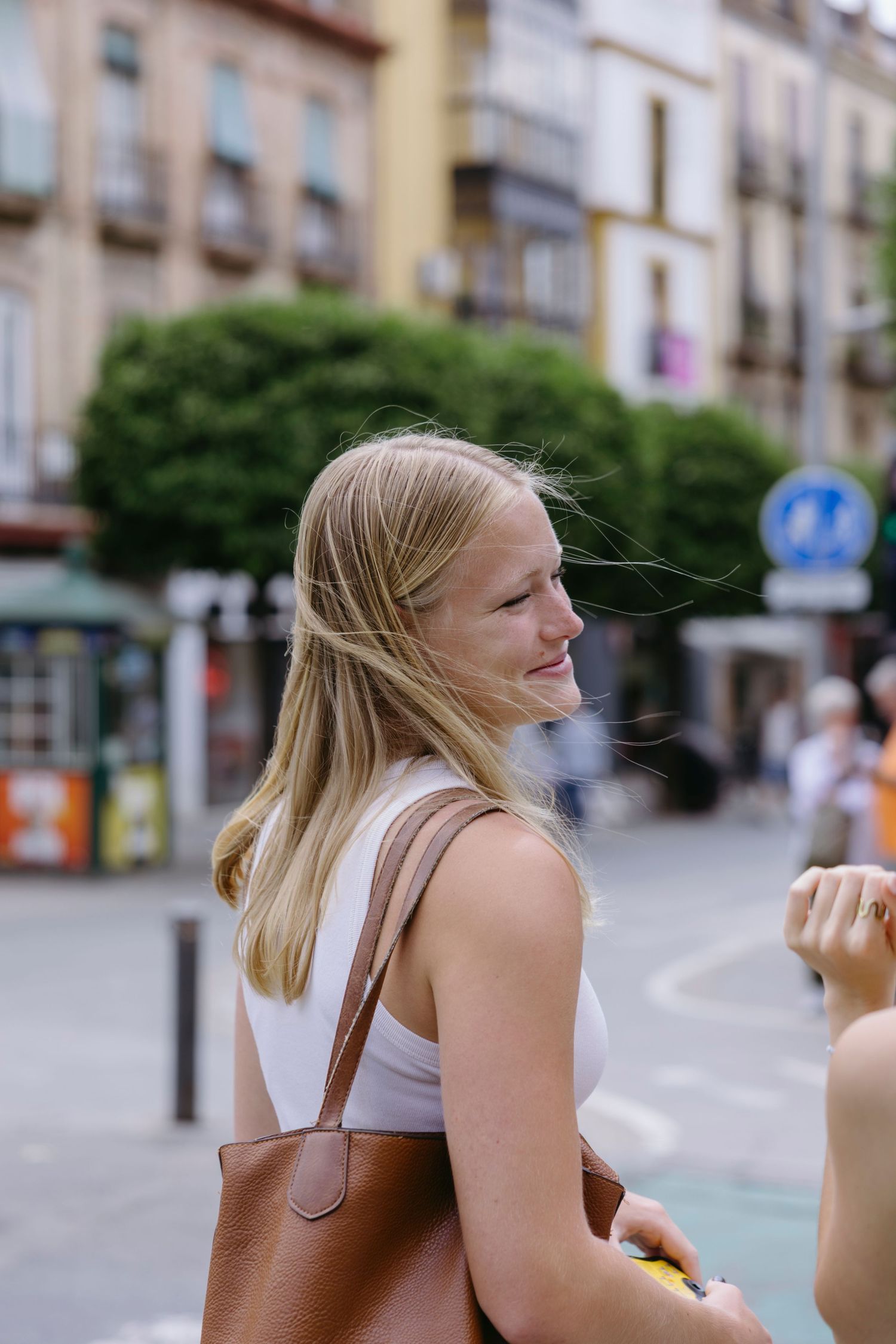 Young person in white top with brown leather bag smiling on a European city street with buildings in background.