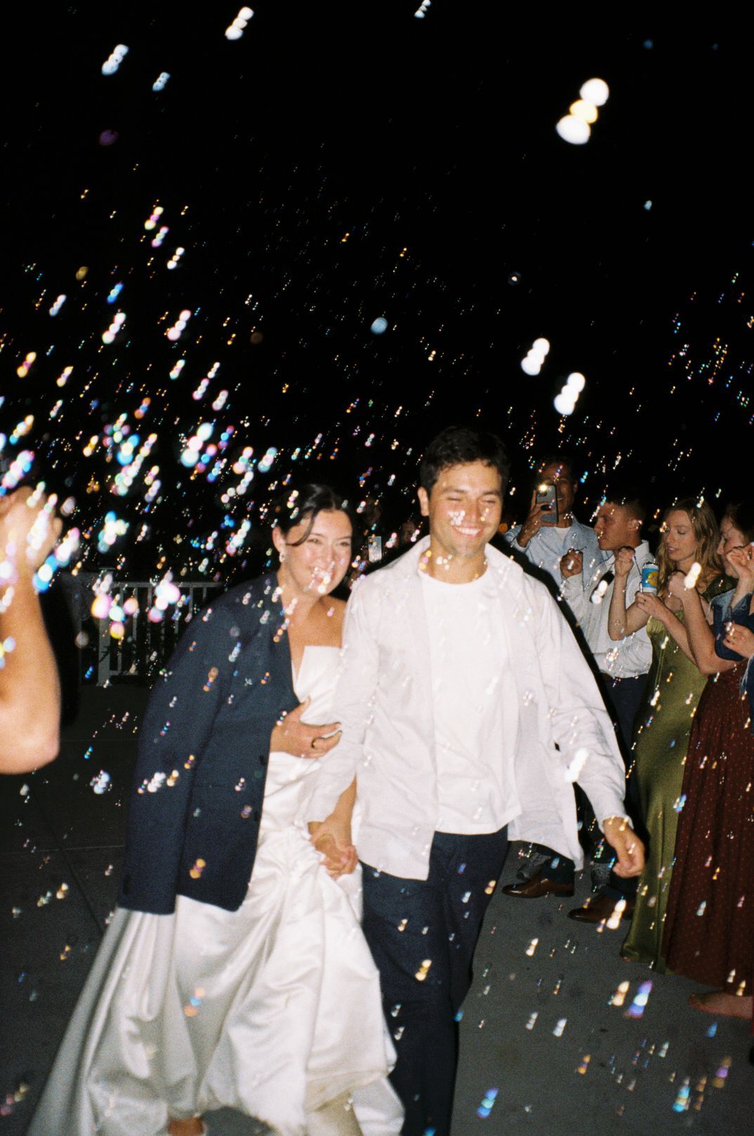 A joyful bride and groom walk through a bubble send-off surrounded by cheering guests at their outdoor evening wedding reception.