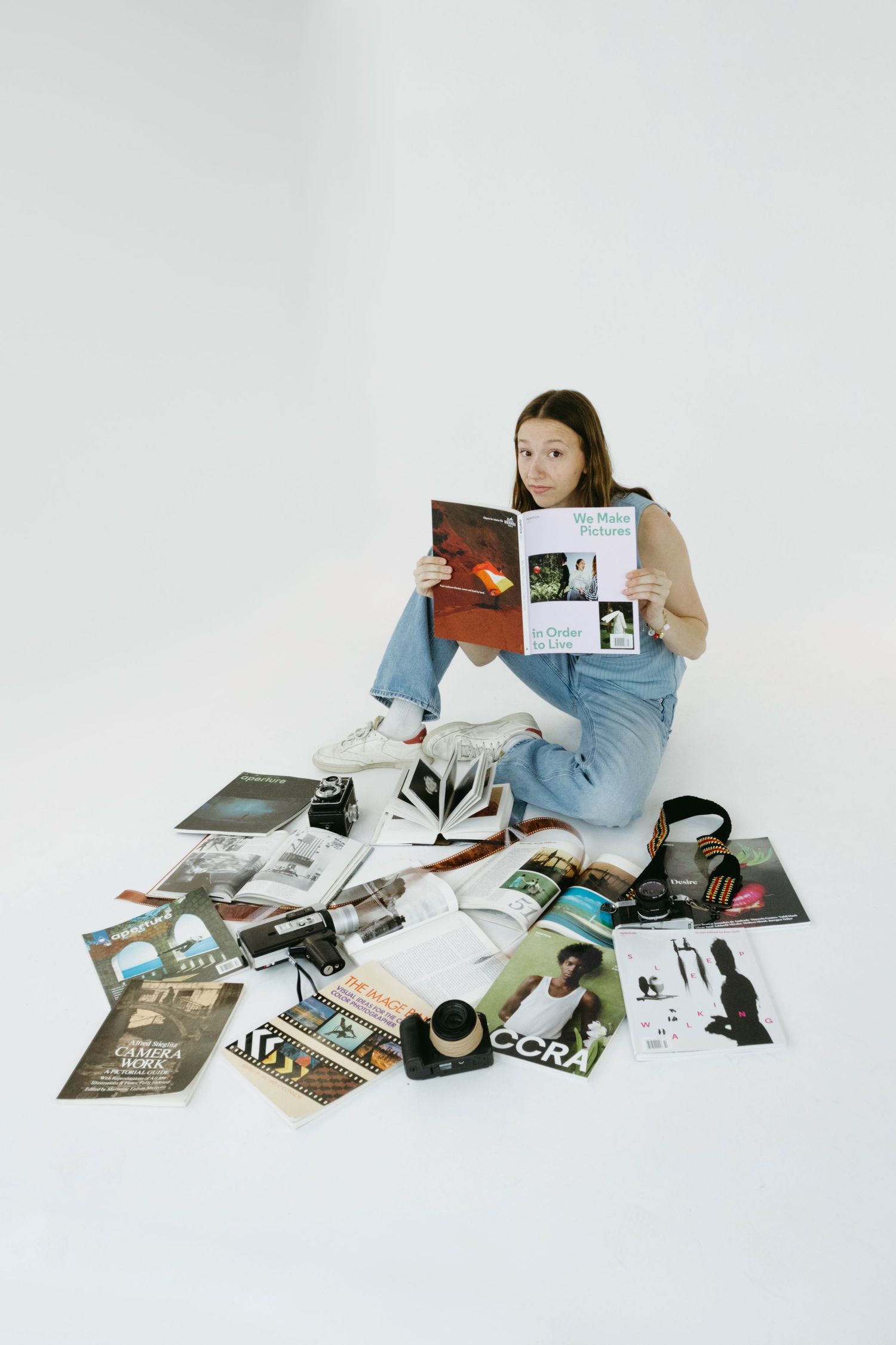 Person in casual denim sits surrounded by scattered magazines, photographs and a coffee cup on white background.