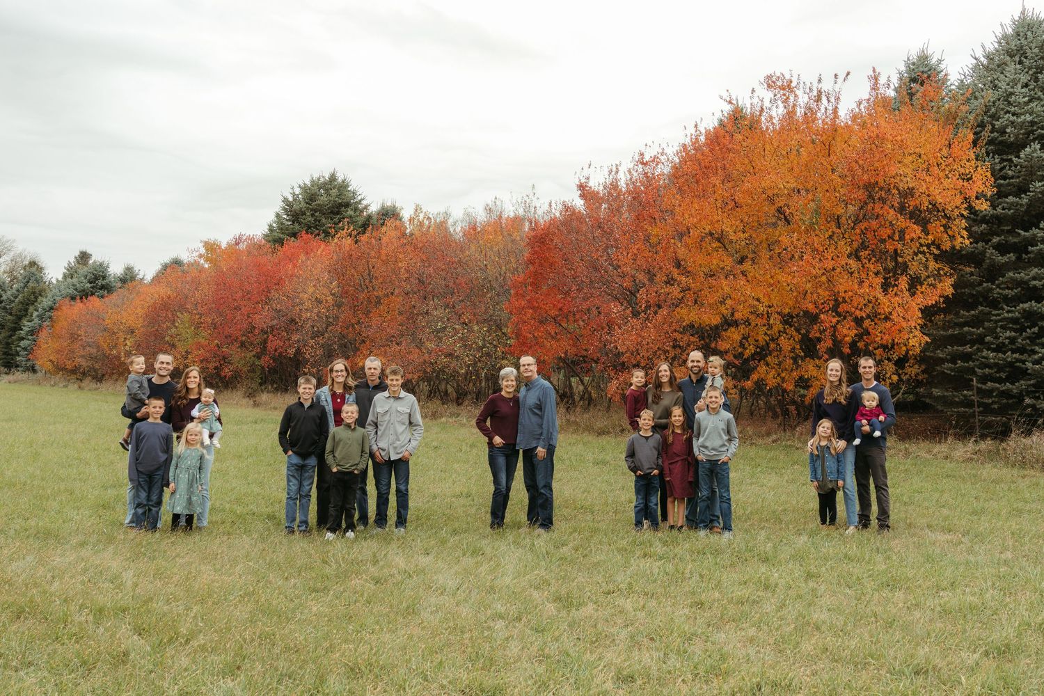 Groups of people stand in a grassy field with vibrant orange and red autumn trees in the background.