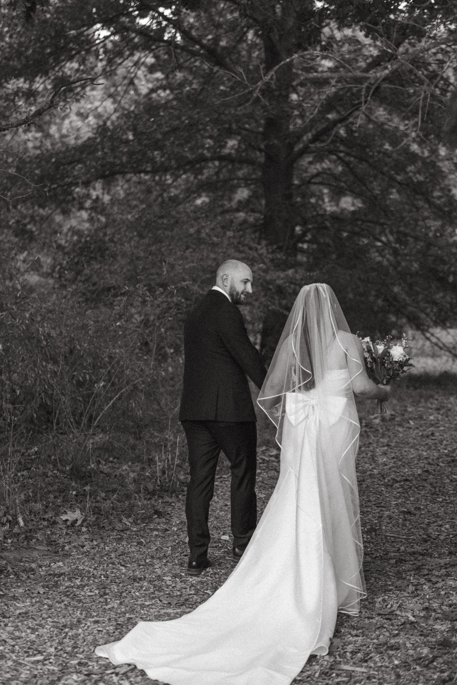 A black and white wedding photo showing a couple in formal attire standing together in a wooded outdoor setting.