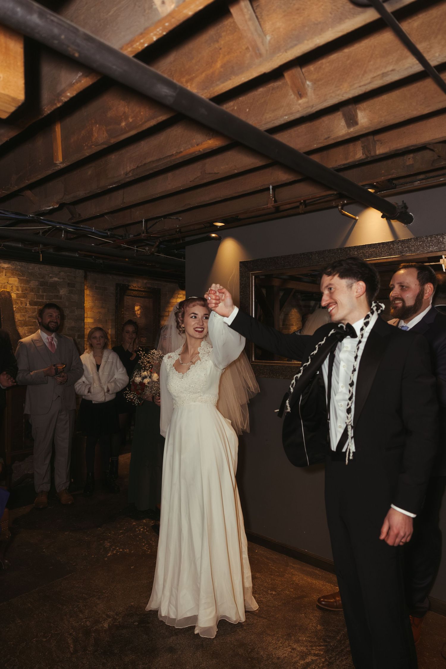 A couple shares a romantic first dance at their wedding reception in a rustic venue with exposed wooden beams.