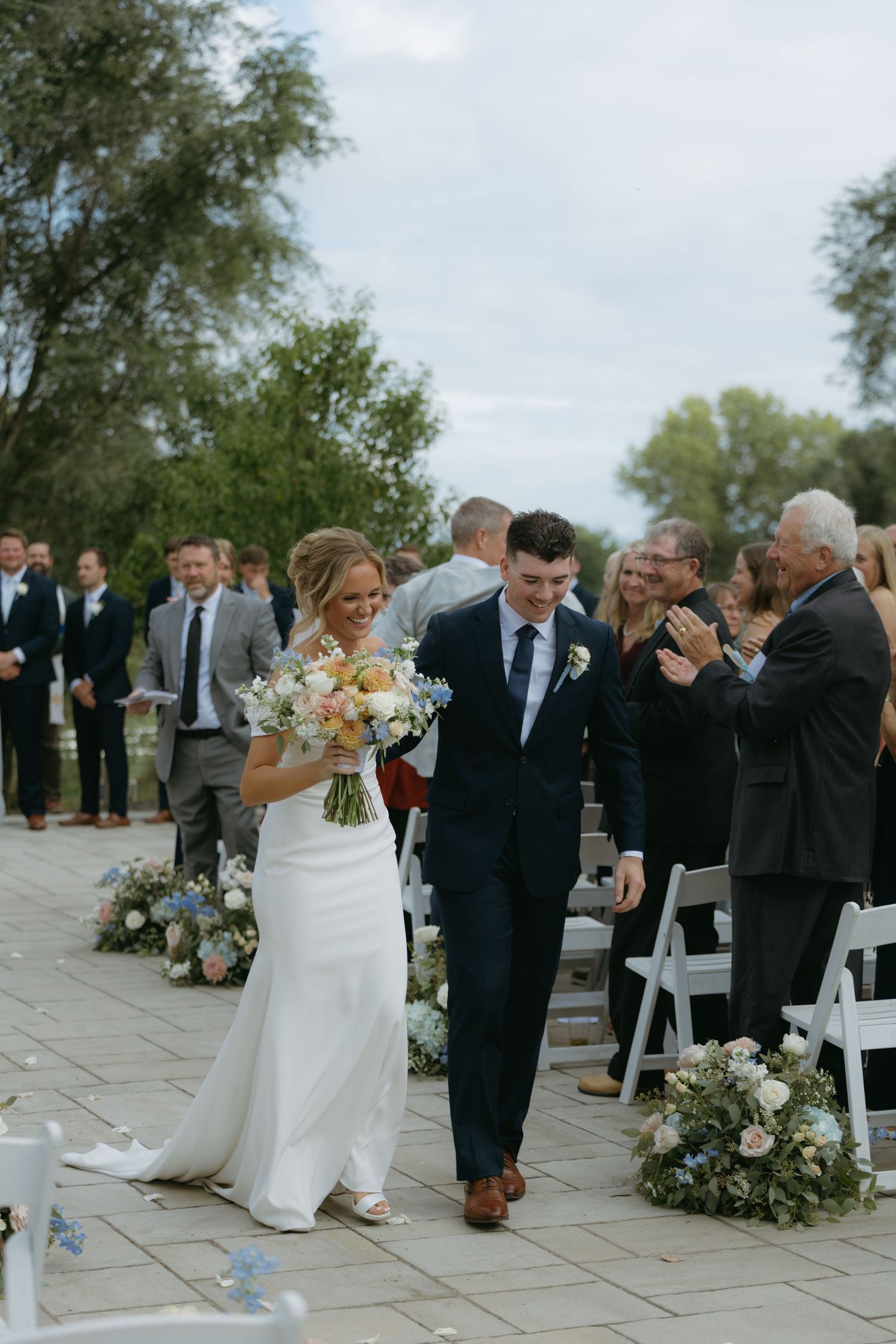Newlyweds walk down the aisle at an outdoor wedding ceremony with guests and floral arrangements on the stone patio.