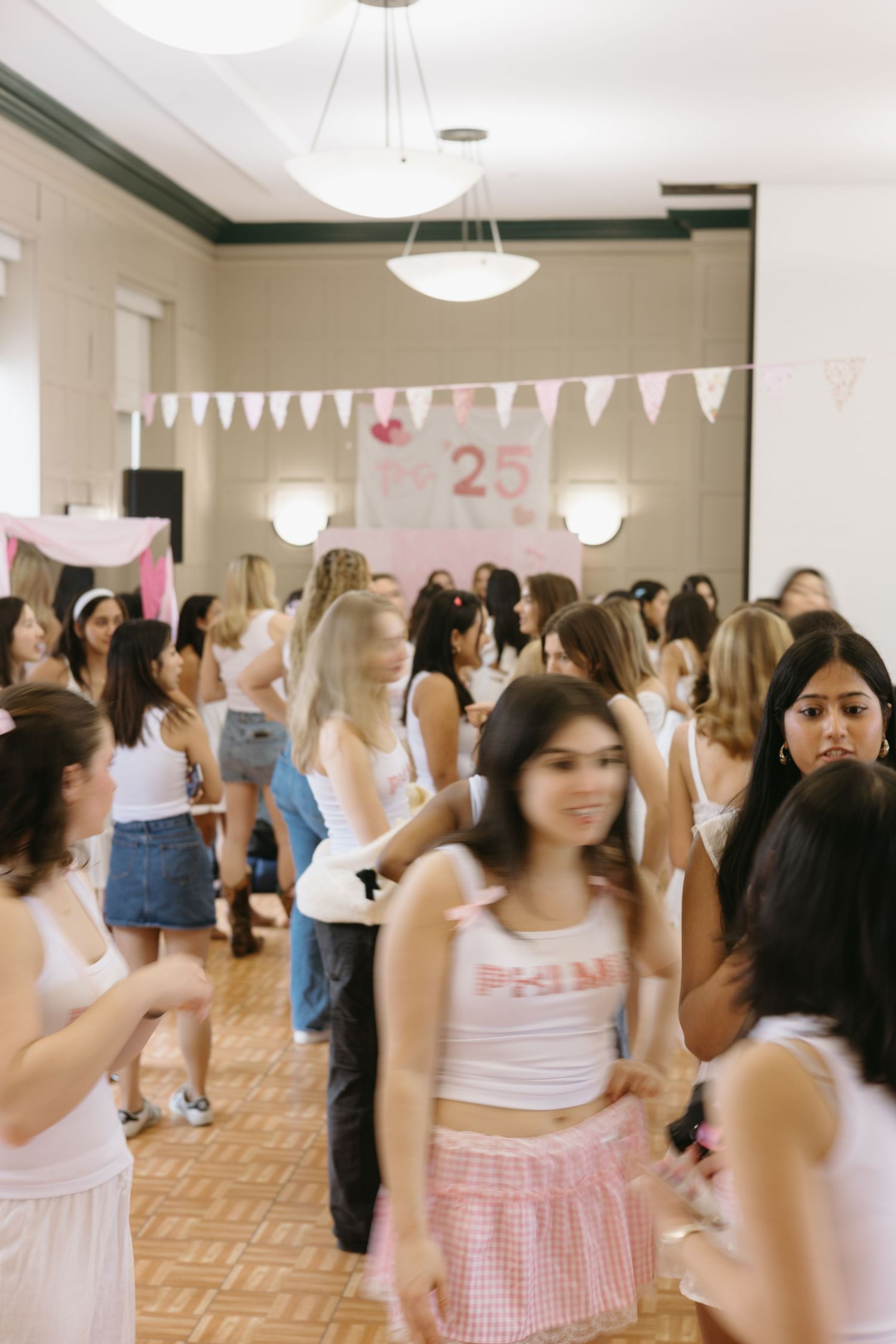 People gather at a party decorated with white pennant banners and a pink '25' sign in an indoor venue with hardwood floors.
