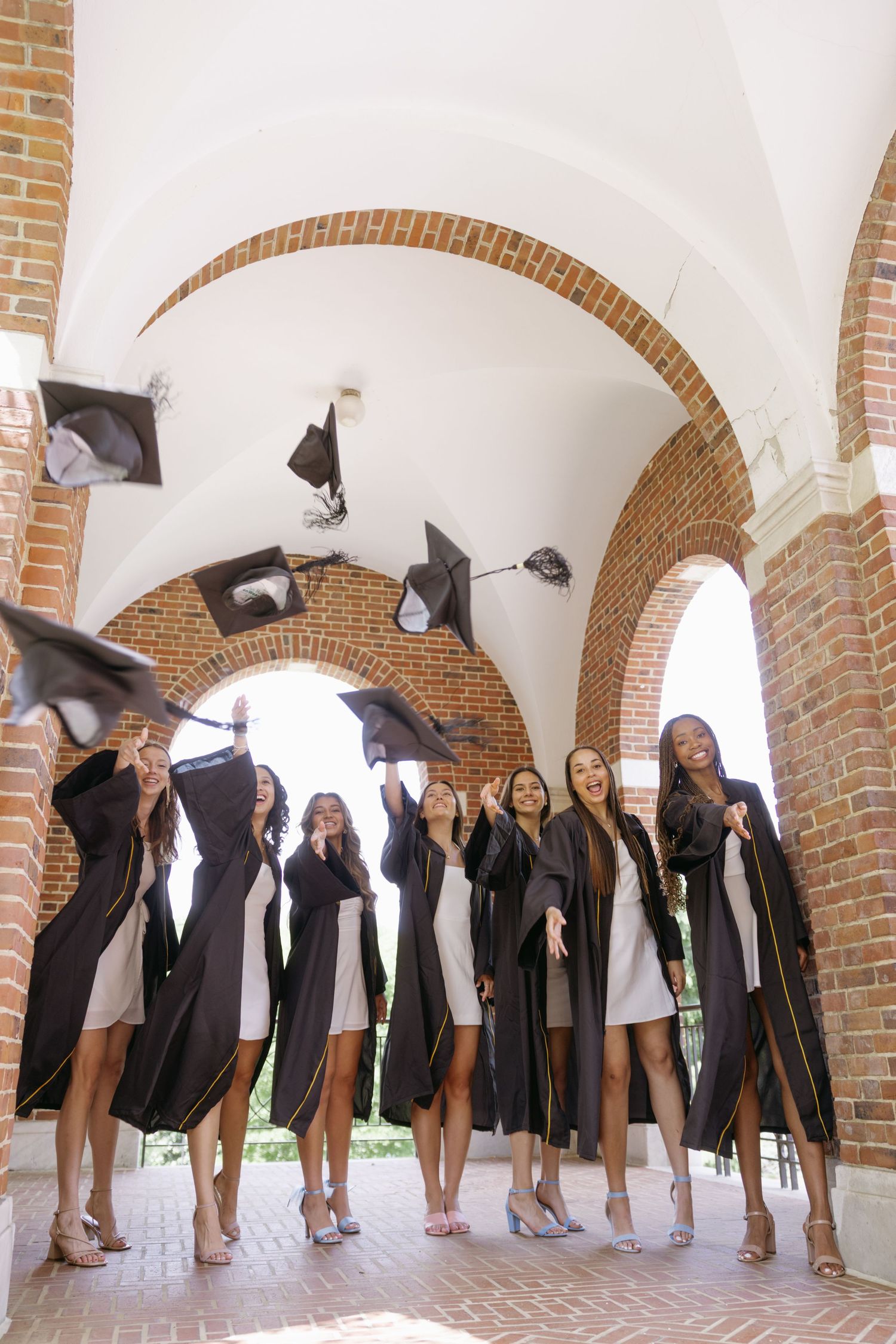 A group of graduates in black gowns toss their graduation caps in an archway walkway with brick columns.