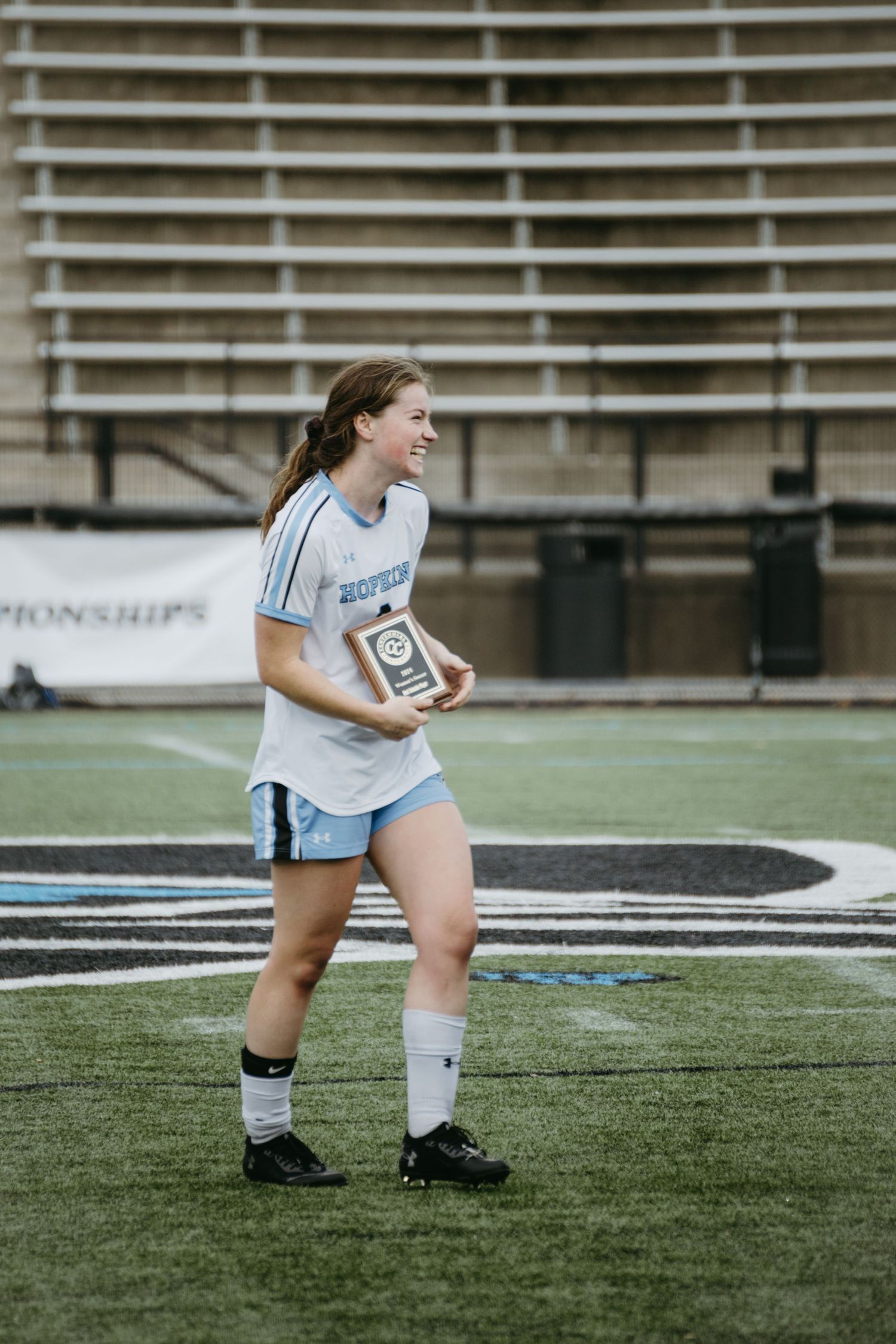 Player holding an award trophy while standing on an athletic field wearing light colored uniform.