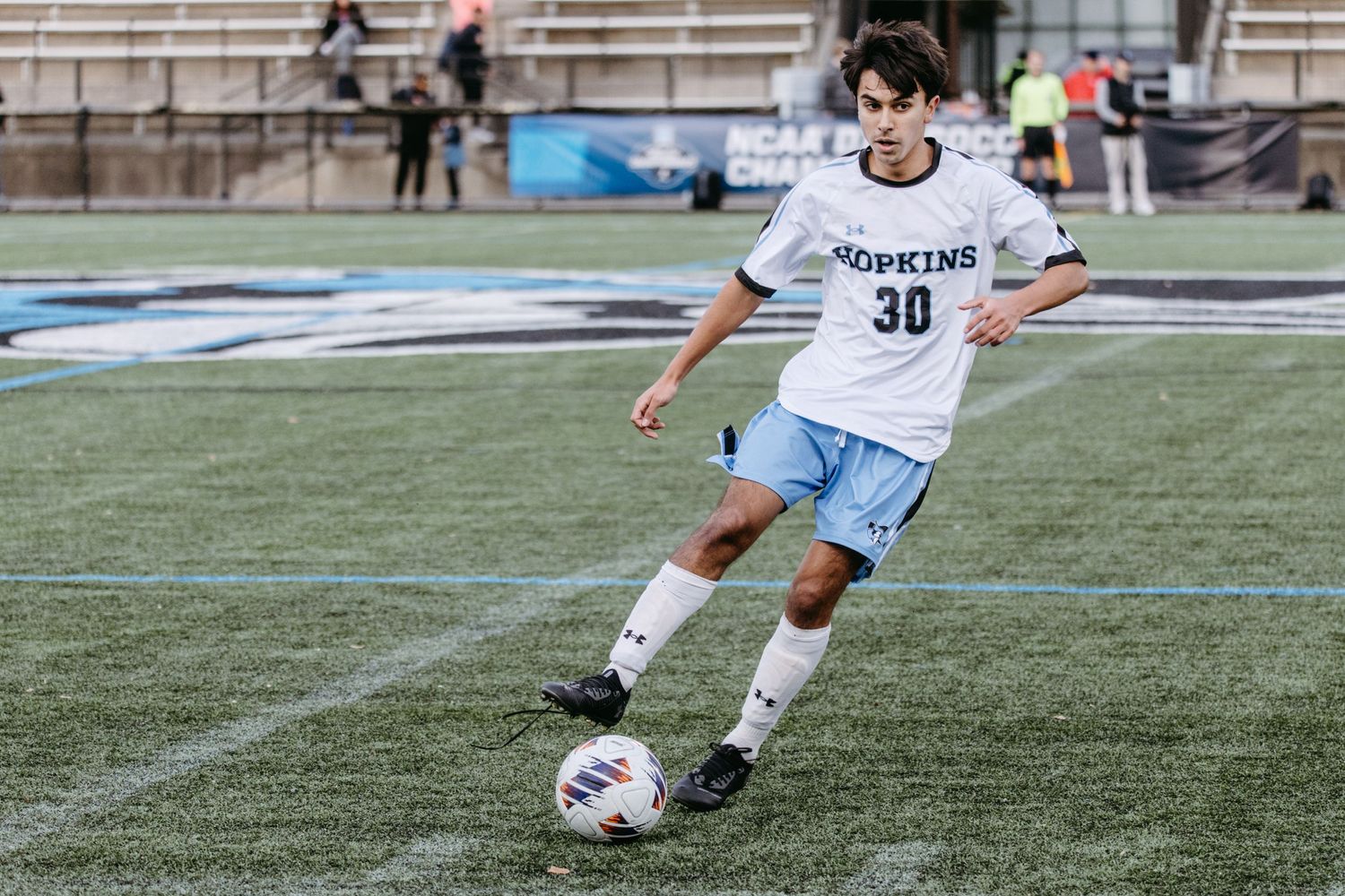 Soccer player in white uniform dribbles ball on artificial turf field.