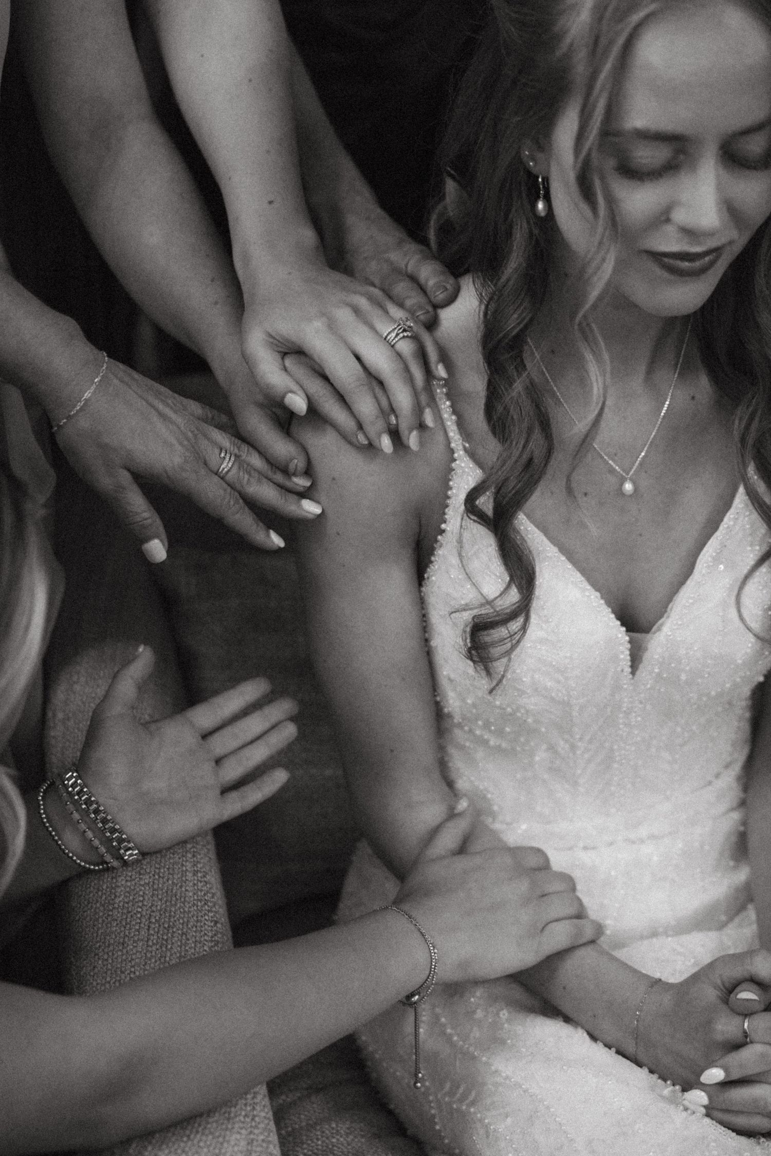 Black and white photo of a bride in a lace gown surrounded by bridesmaids holding hands in a prayer circle.